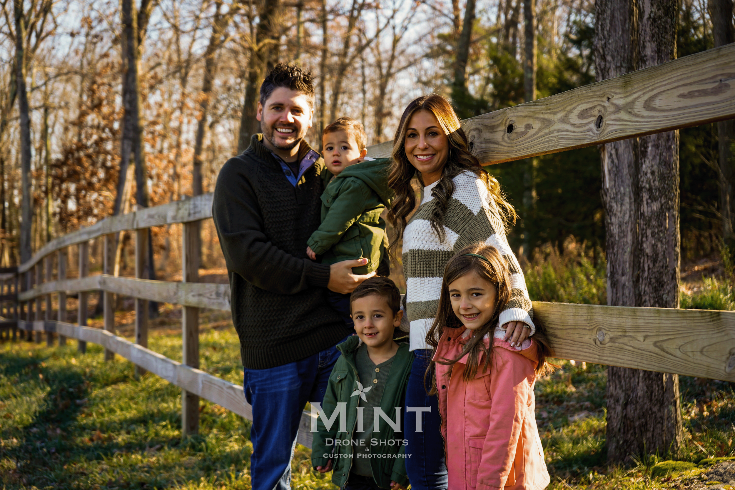 A family of five standing together outdoors next to a wooden fence in a forested area. They are smiling, with the parents holding a toddler. The scene is lit by a warm, natural light.