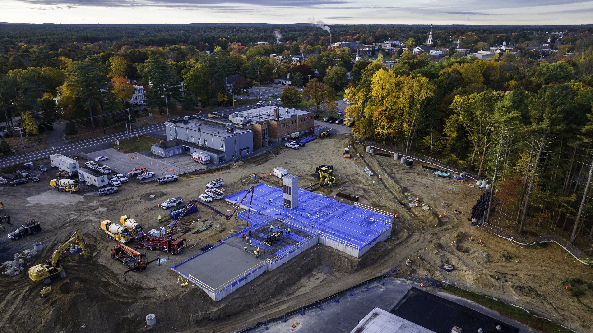 Aerial view of a construction site with machinery and vehicles, surrounded by trees in autumn colors. A building foundation is visible, and a small town with church steeples is in the background.