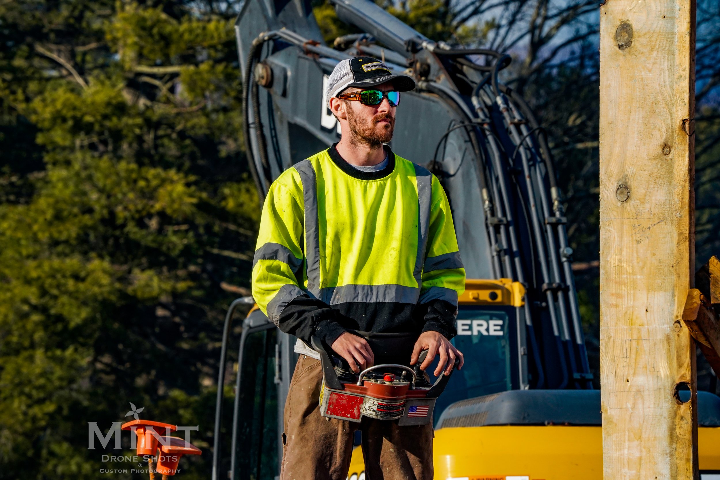 Construction worker in high-visibility clothing operating machinery with a remote controller, standing in front of excavator on a job site.