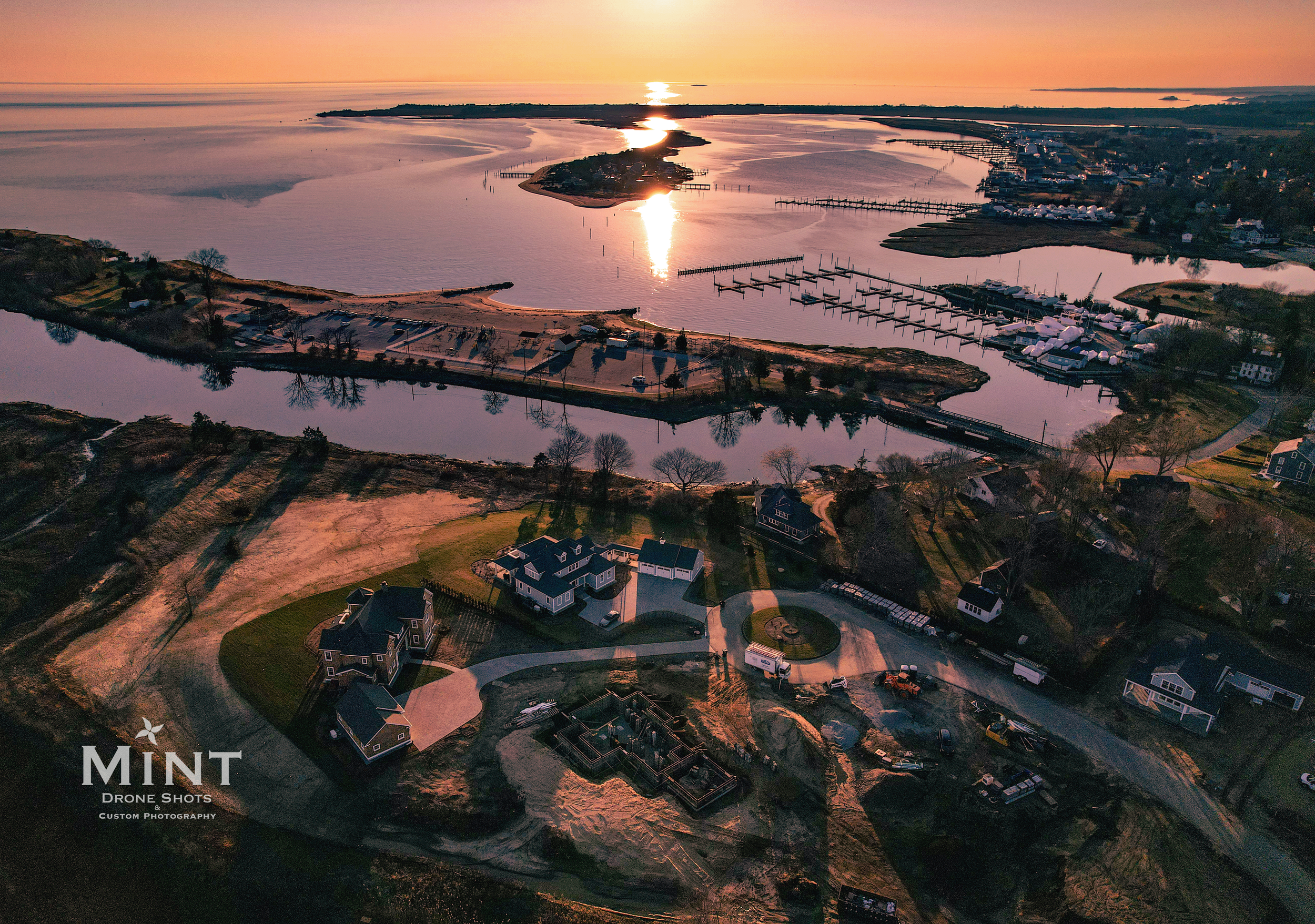 Aerial view of a coastal area at sunset, featuring a calm river, docked boats, residential homes, and an illuminated horizon.