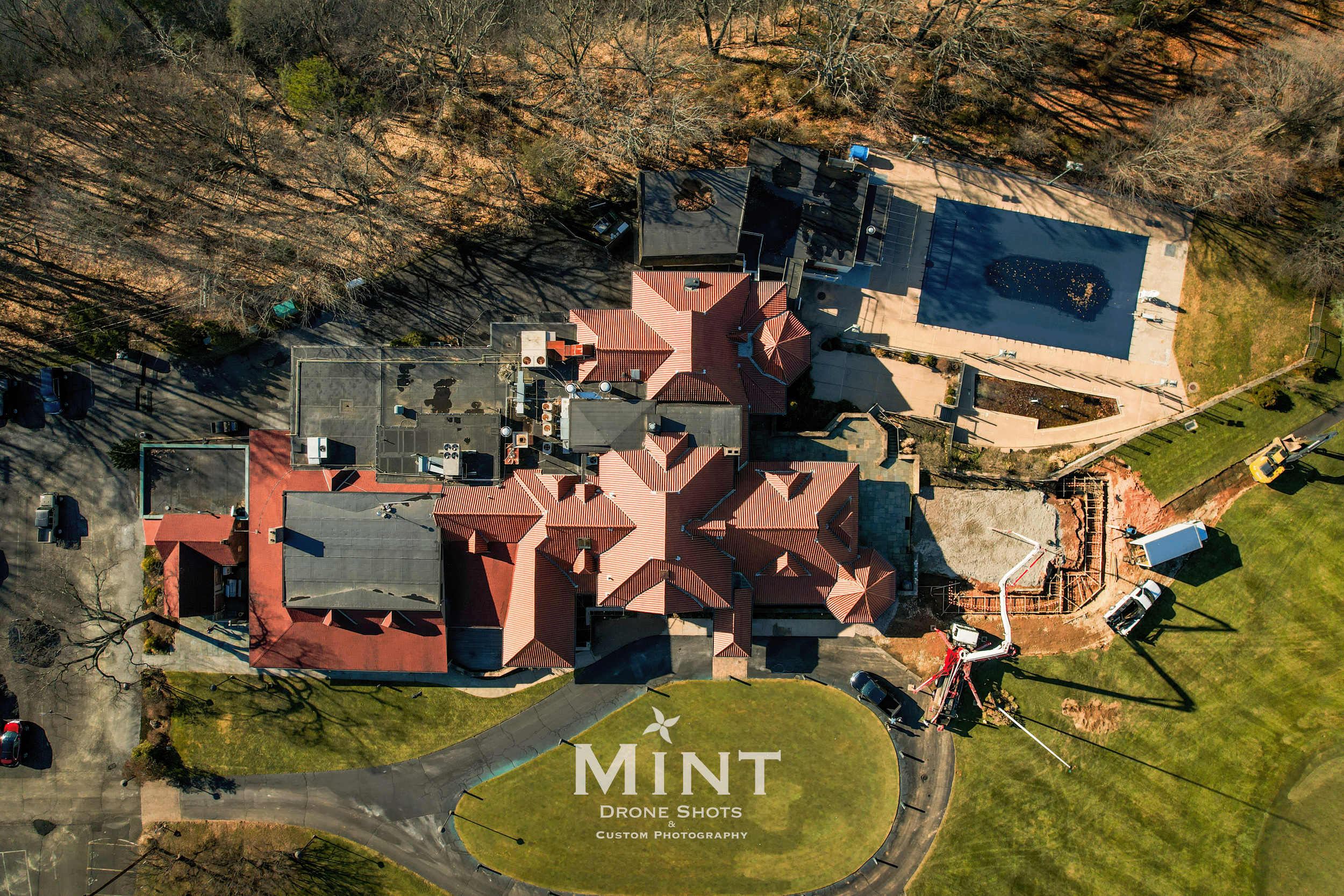 Aerial view of a large estate featuring a building with multiple red roofs, surrounding trees, a swimming pool, and vehicles. The image includes construction equipment and a circular driveway. The scene is labeled "Mint Drone Shots: Custom Photograph