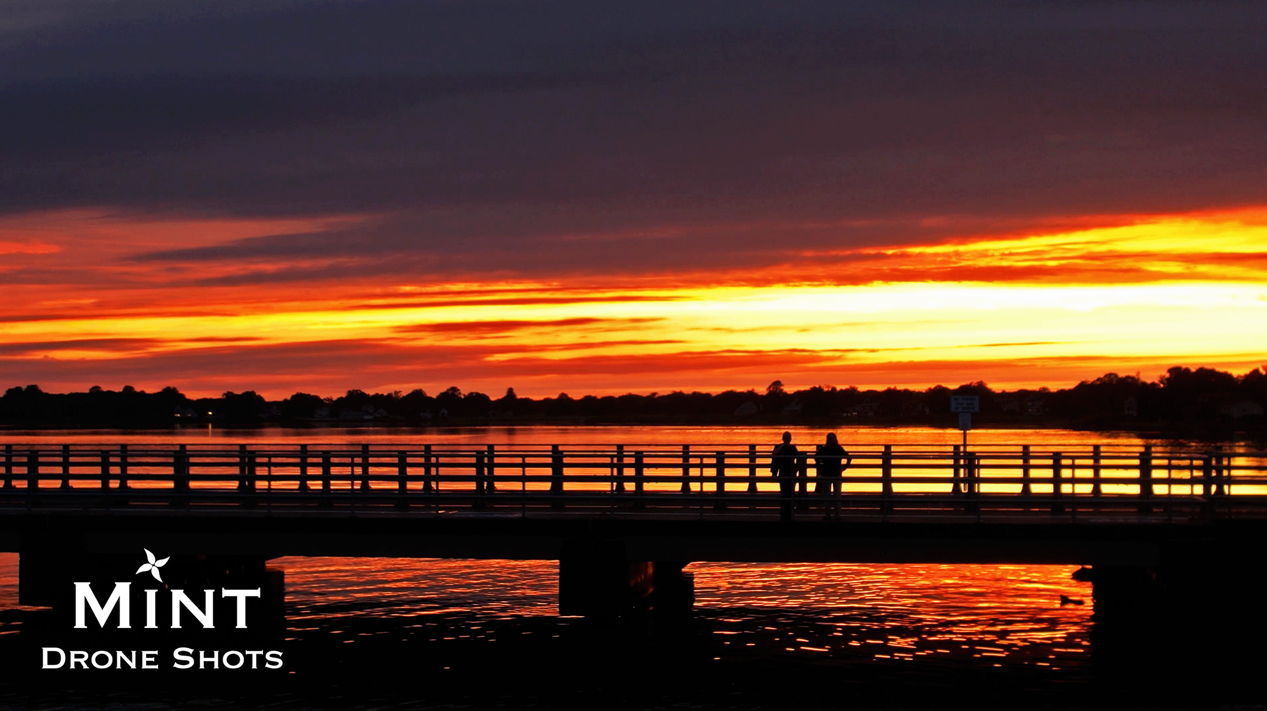 Silhouette of a bridge against a vibrant orange sunset over a body of water, with two people standing on the bridge.