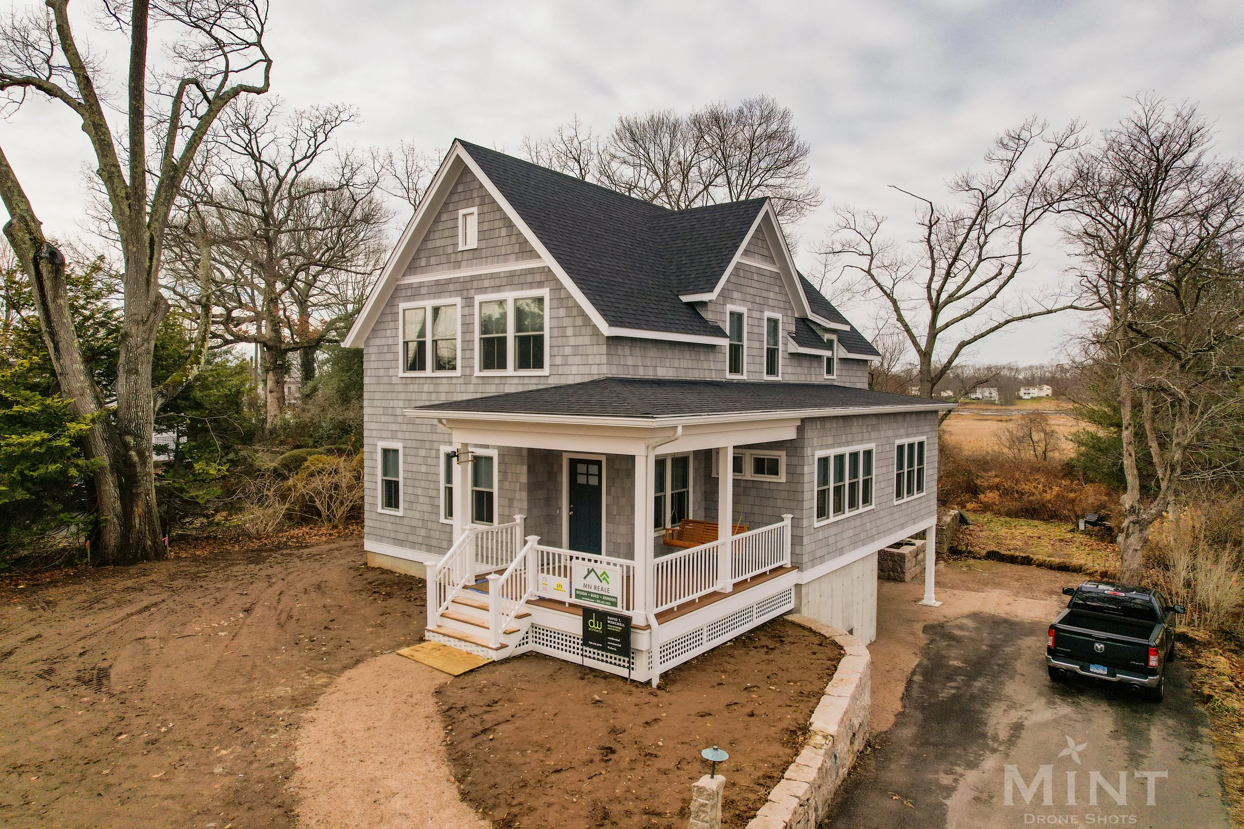 Newly constructed gray shingle-style house with a black roof, surrounded by bare trees and a driveway with a parked black truck. A porch with white railings is visible.