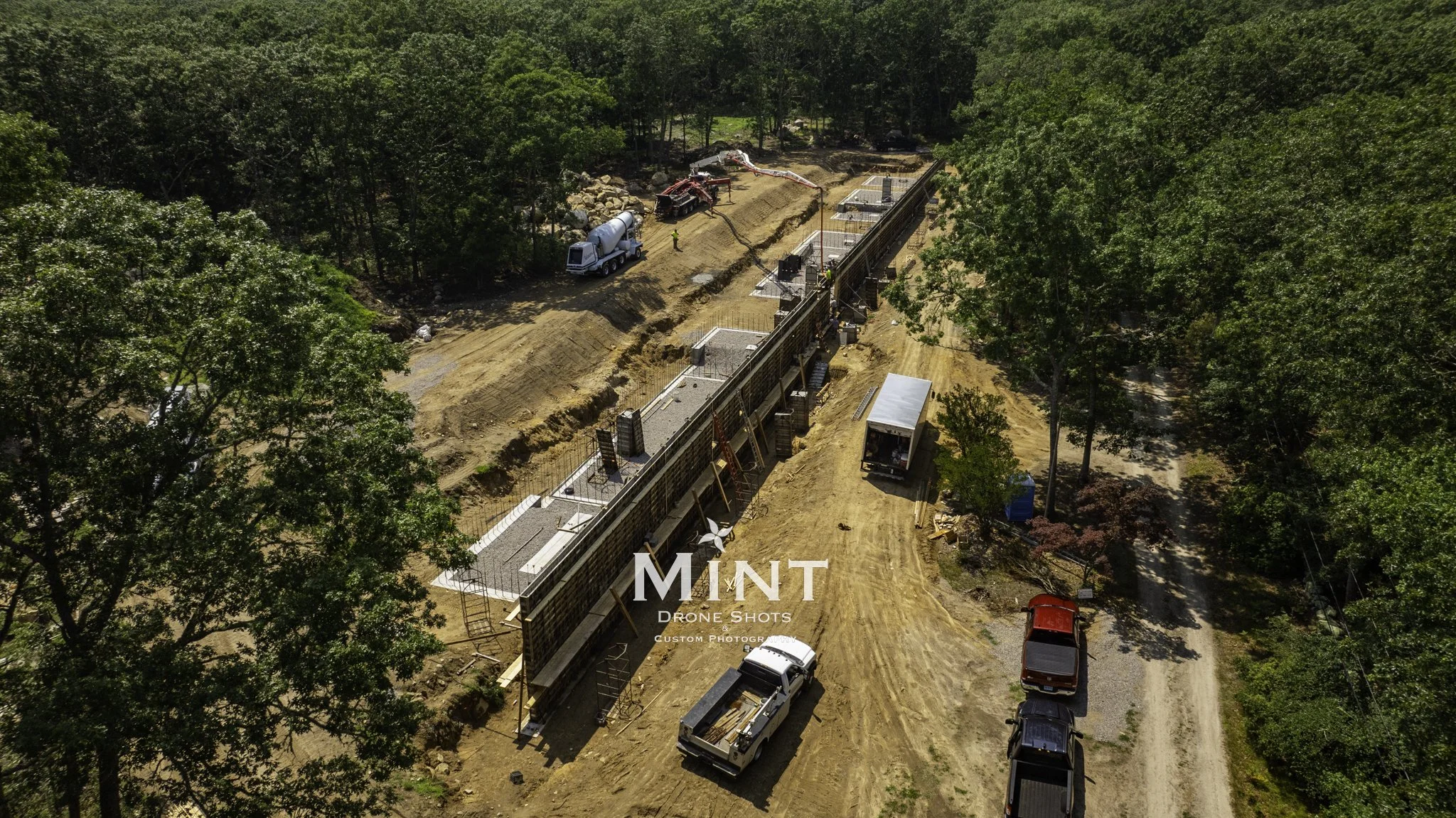 Aerial view of a construction site with trucks, unfinished concrete structures, and surrounding forest.