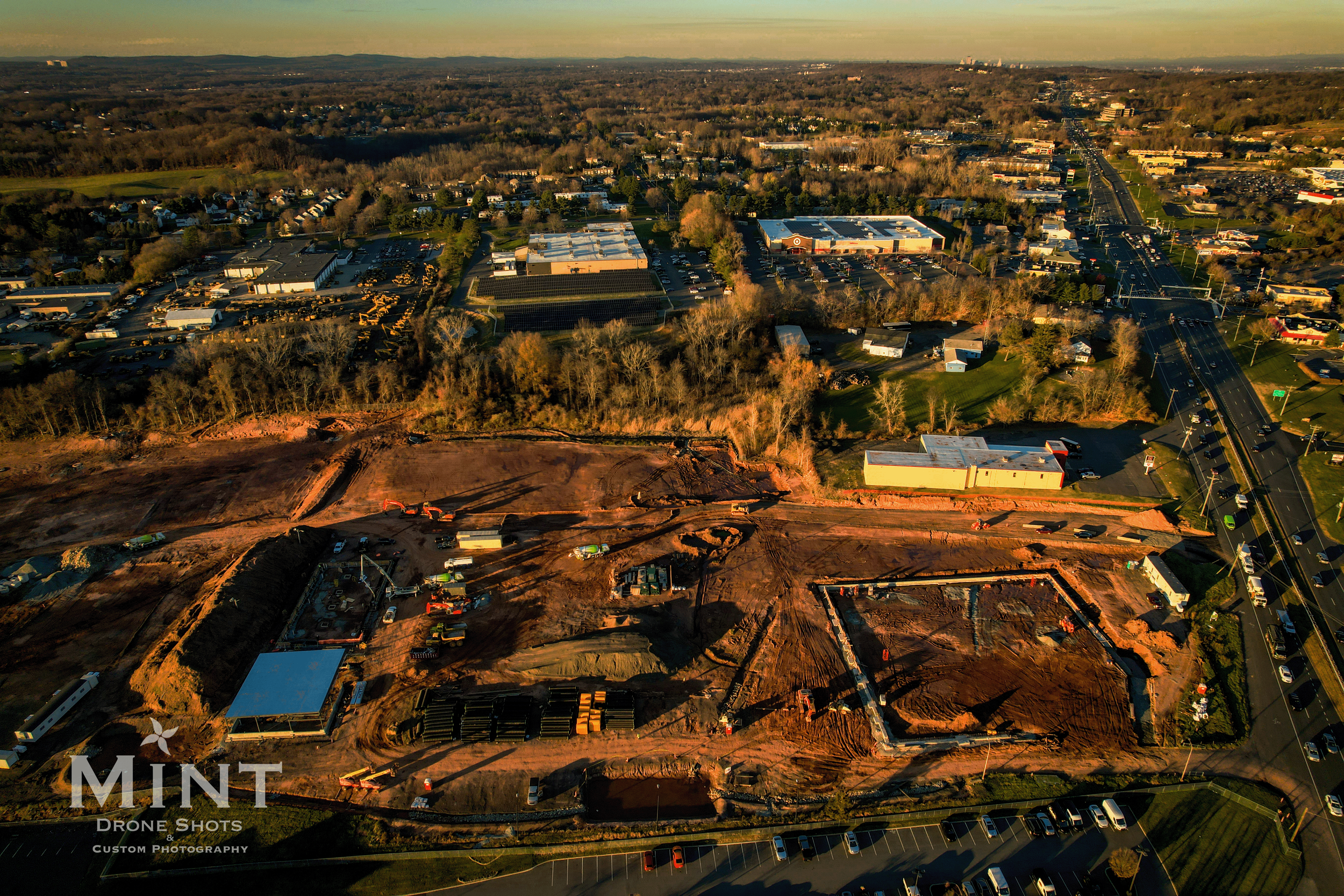 Aerial view of a construction site with surrounding landscape, buildings, and a road.
