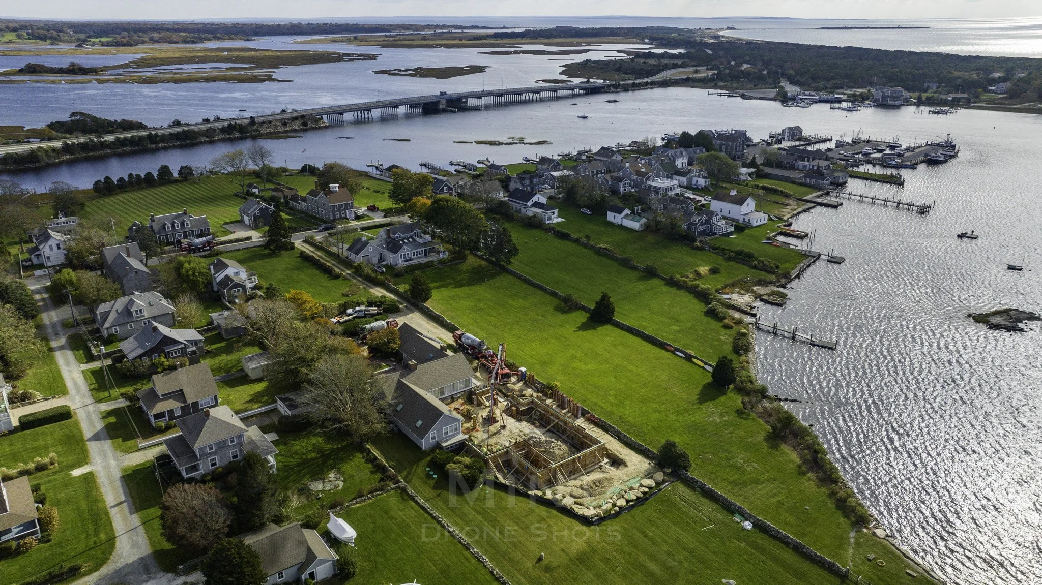 Aerial view of a coastal residential area with houses, green lawns, a construction site, and a river with a bridge and docks.