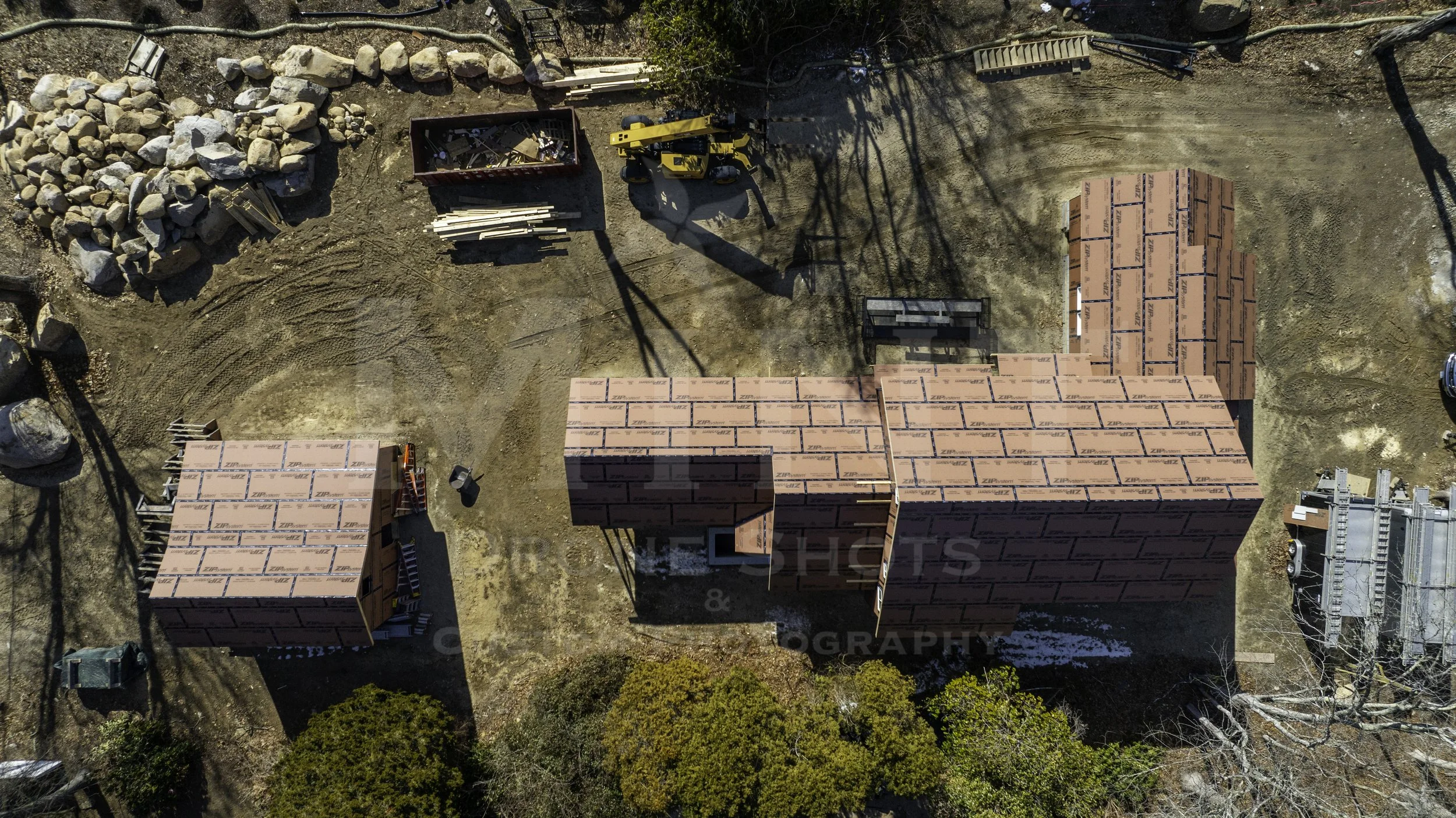 Aerial view of a construction site with partially built structures and construction materials, including a stack of wood and a bulldozer. The structures have roofing making progress.