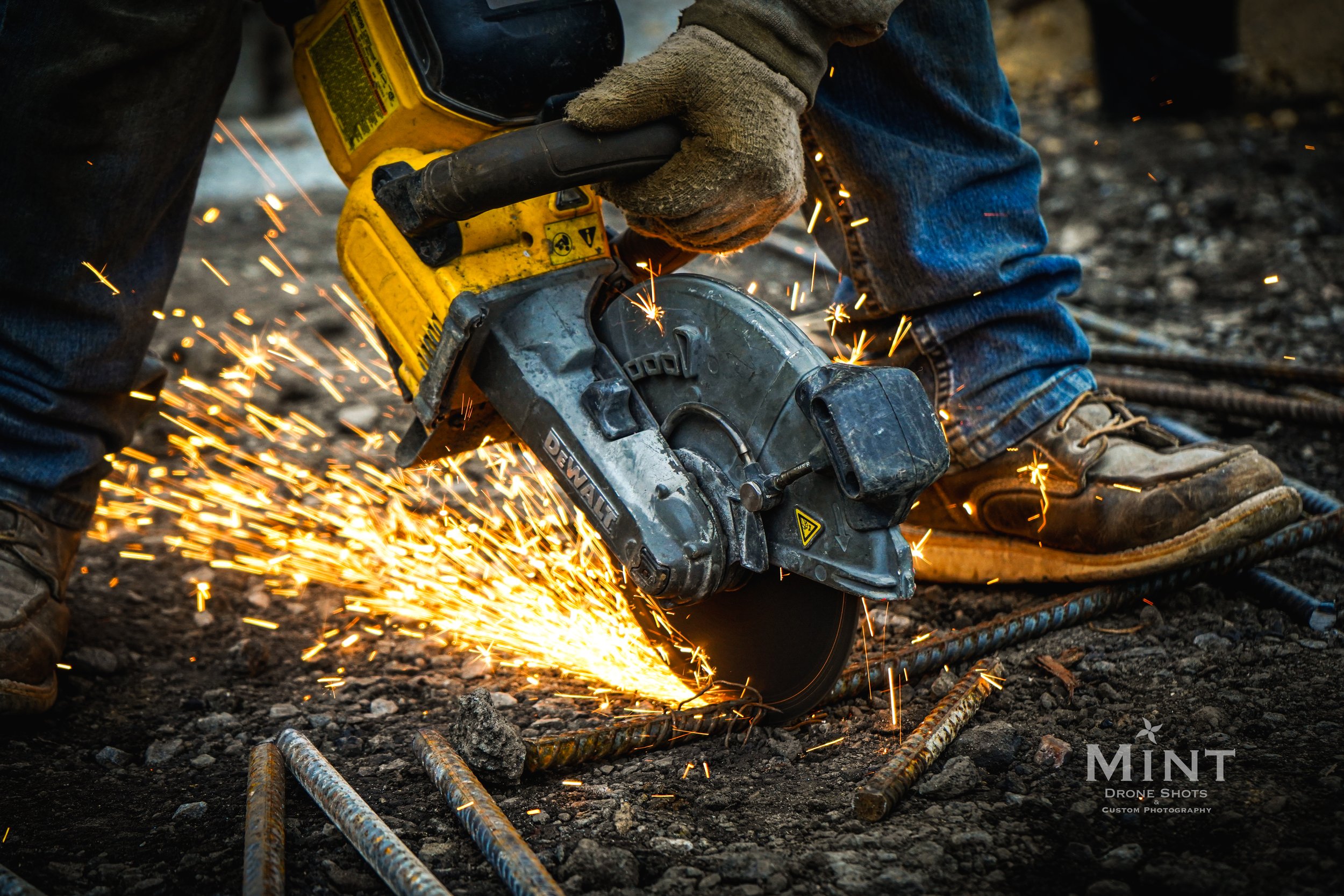 Construction worker using a circular saw to cut metal rods, with sparks flying, wearing protective gloves and boots.