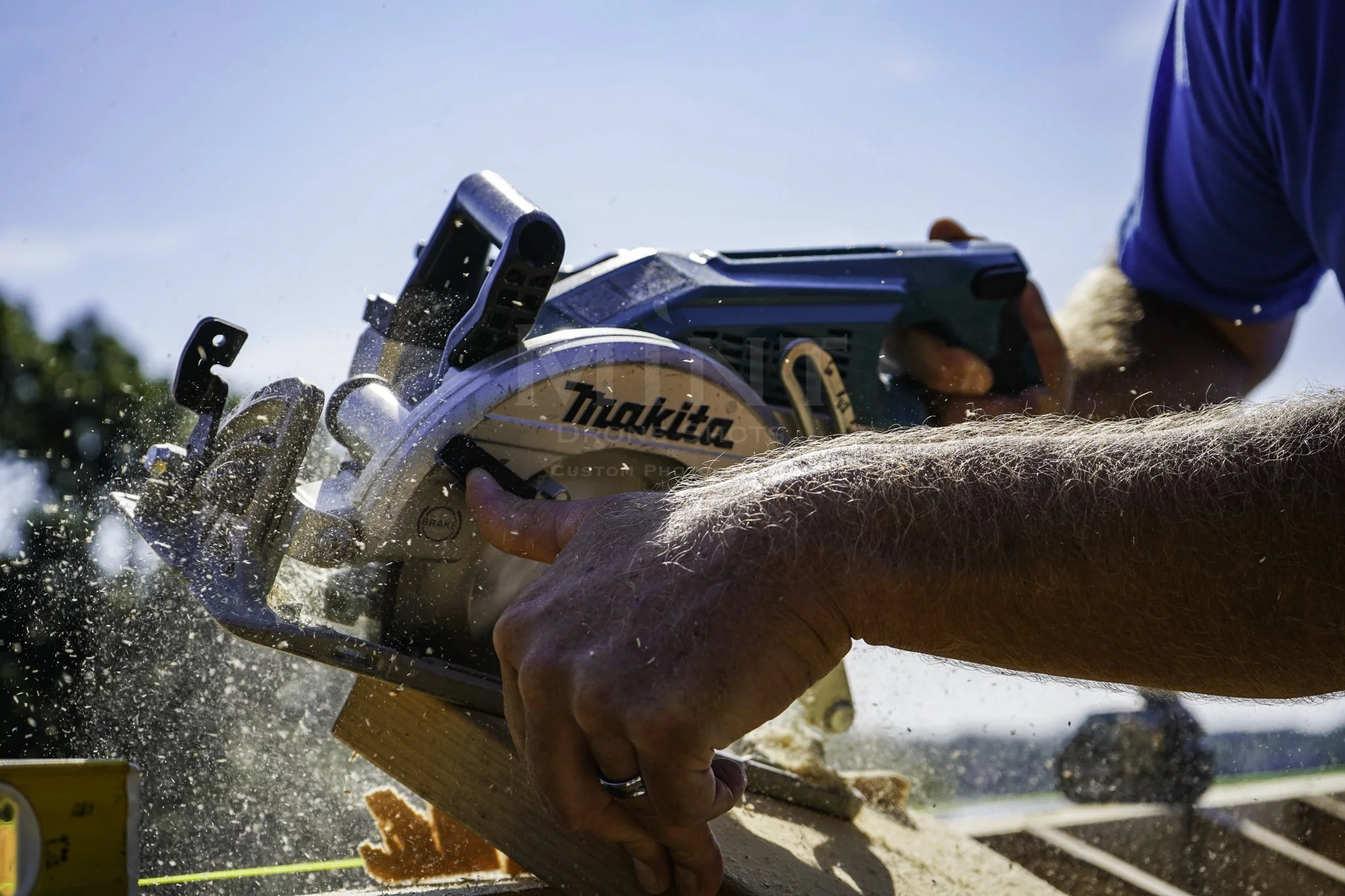 Person using a circular saw to cut wood, with sawdust flying, under a clear blue sky.