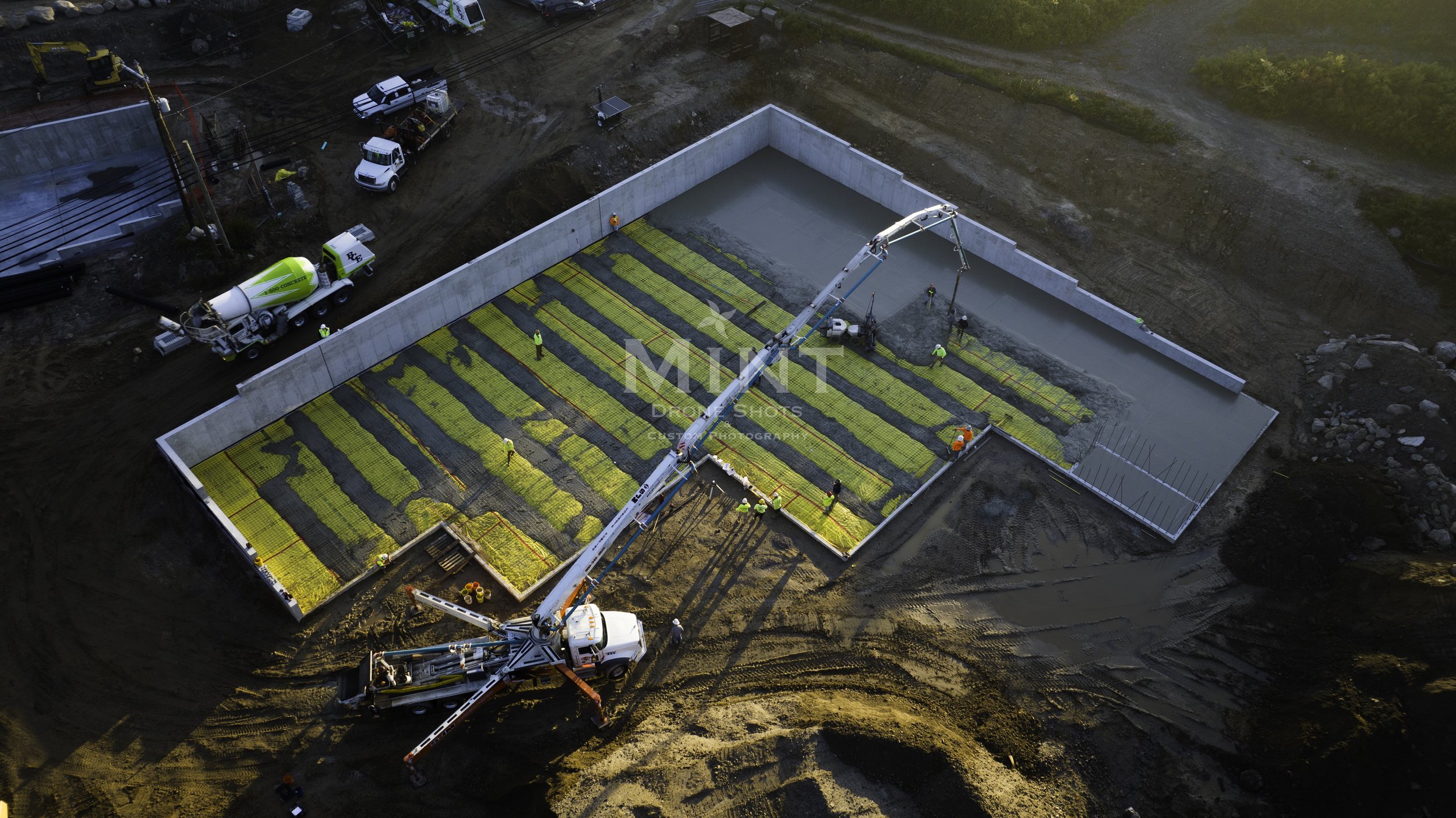 Aerial view of a construction site with concrete pouring equipment and workers. The site features a large rectangular foundation with concrete being poured inside bordered by retaining walls. Several trucks and construction machinery are present.