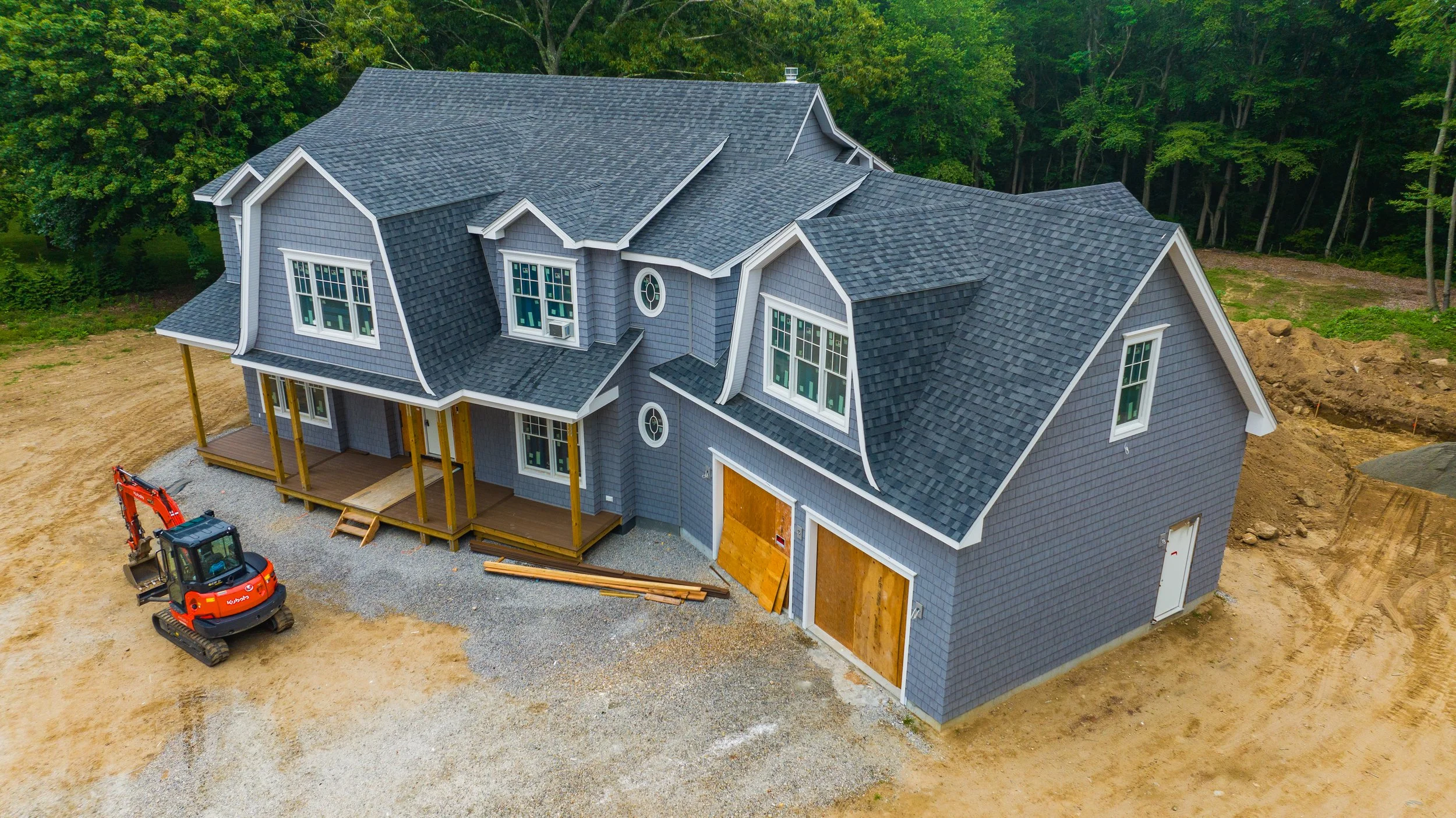 Aerial view of a partially completed two-story gray house with a complex roof, a front porch, and construction equipment nearby.