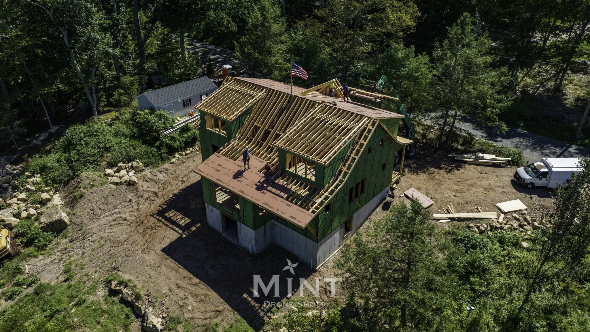 Aerial view of a house under construction with workers on the roof. The building has a partially completed roof and wooden framework. Surrounding the house is a wooded area and a dirt construction site with a white van parked nearby.