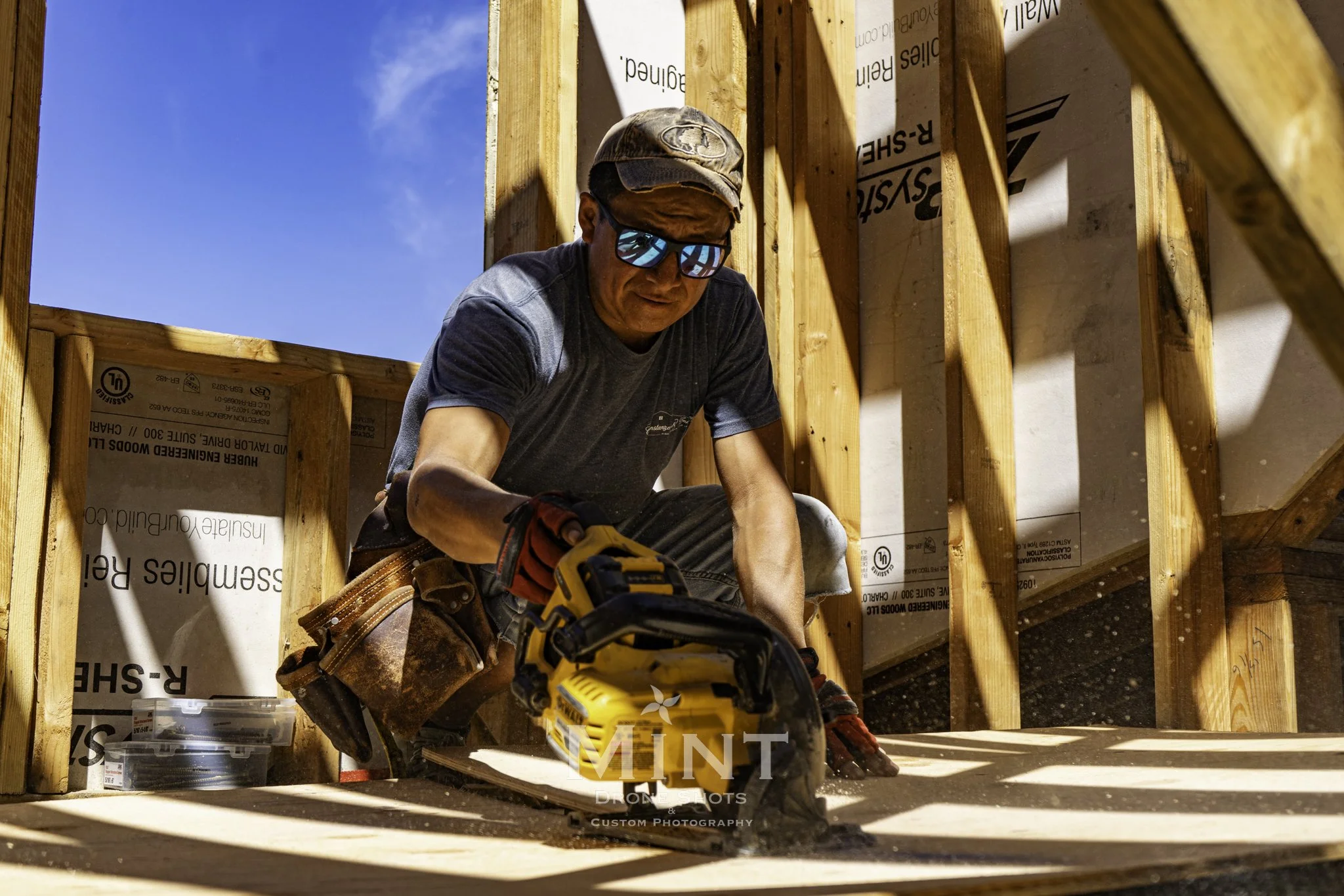 Carpenter wearing sunglasses and a cap using a circular saw in a building under construction with wooden beams and blue sky visible.