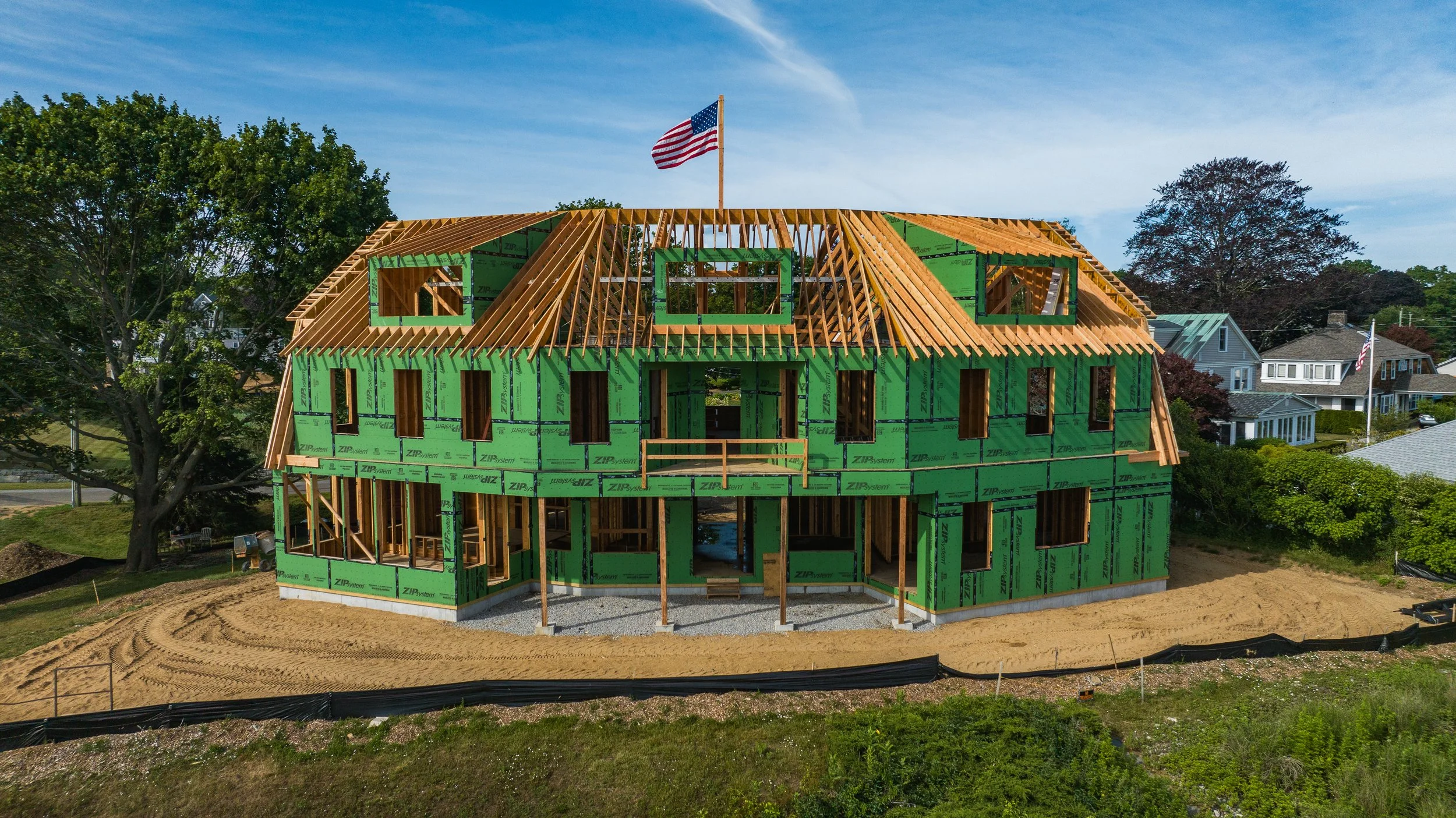 Partially constructed house with green insulation sheathing and wooden framing, featuring an American flag on top; surrounding trees and neighboring houses in the background.
