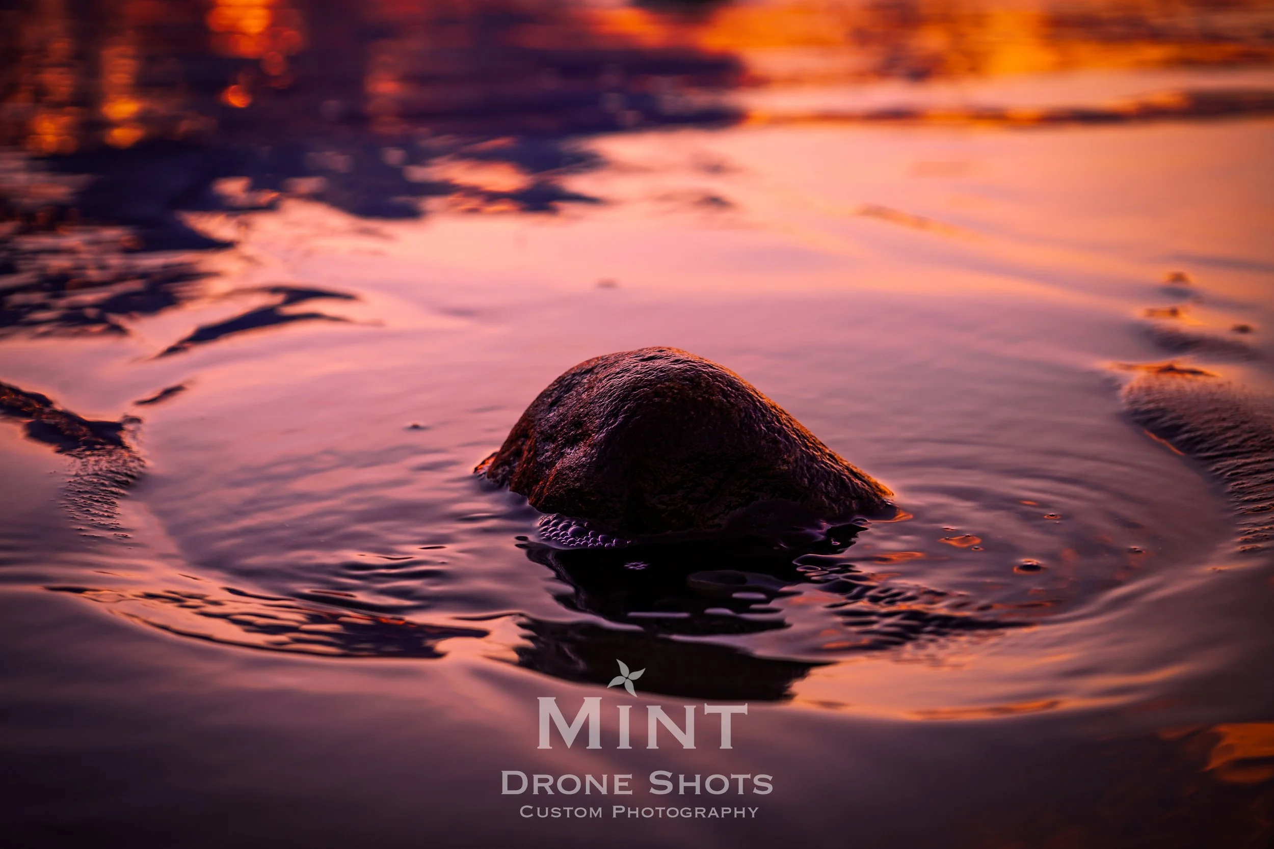 A rock partially submerged in calm water during a sunset, with warm orange and pink reflections on the surface.