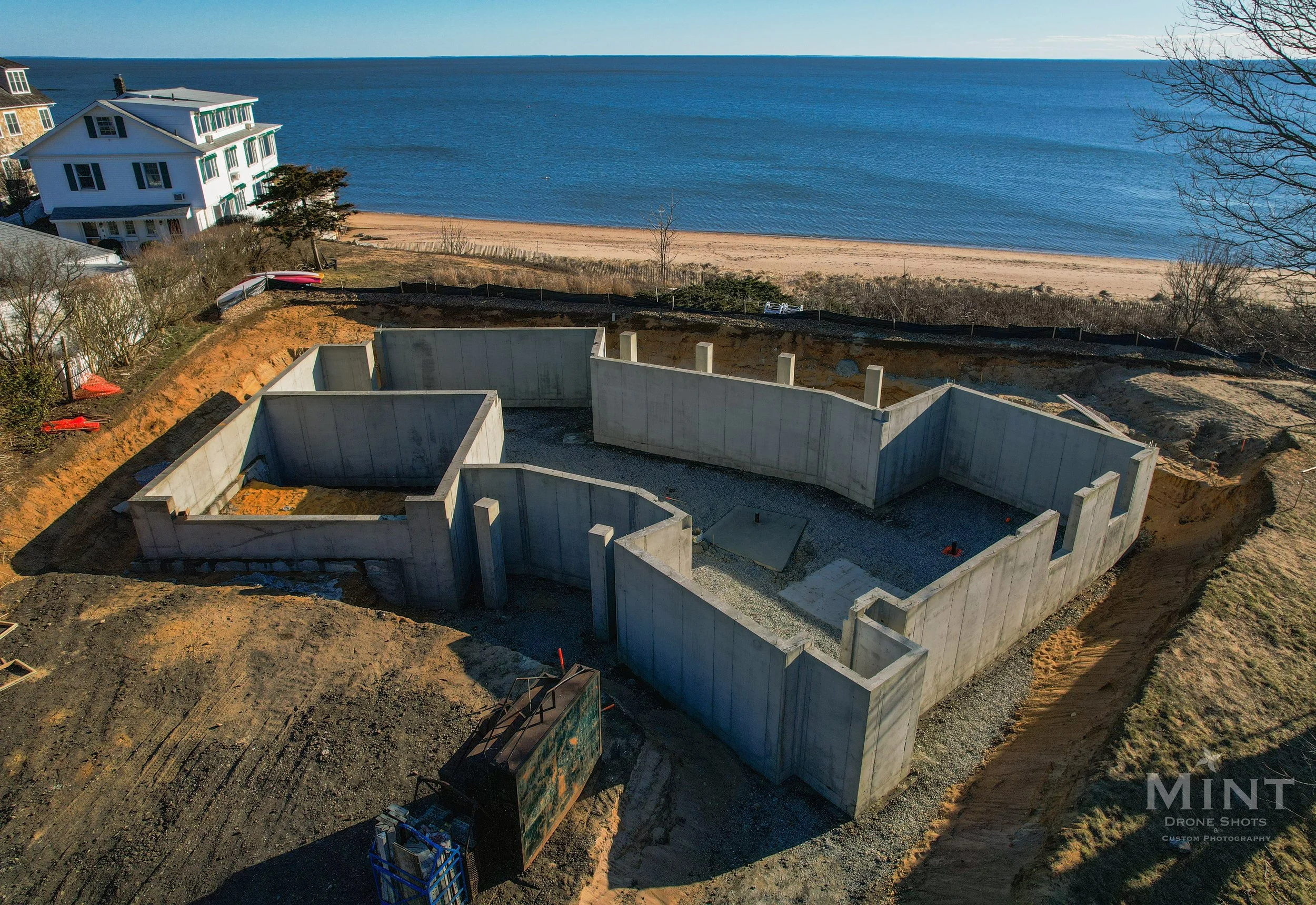 Aerial view of a building foundation under construction near a beach, with adjacent houses and a clear blue ocean in the background.