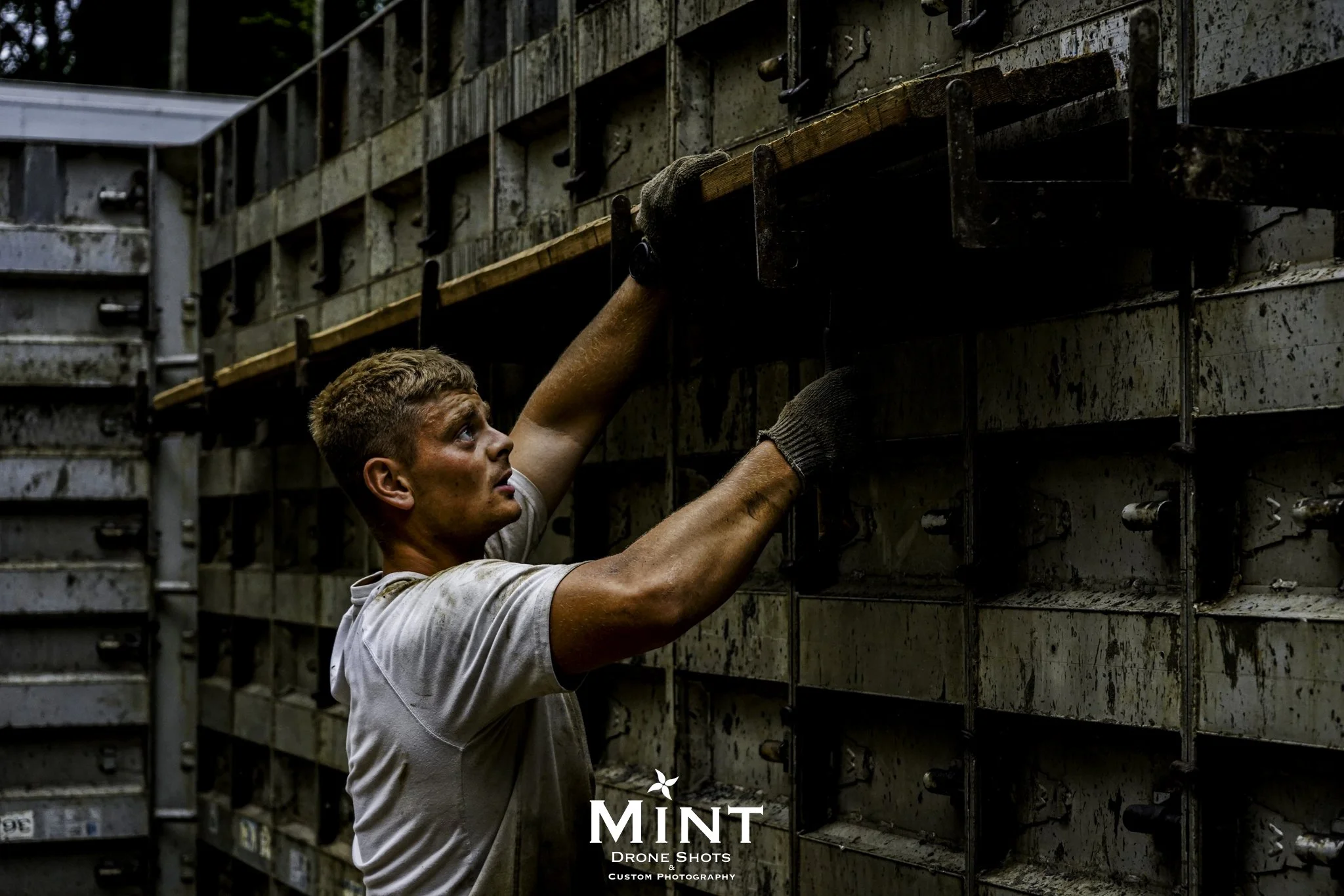 Worker adjusting industrial machinery, wearing gloves and a white shirt, standing next to a large, metal structure.