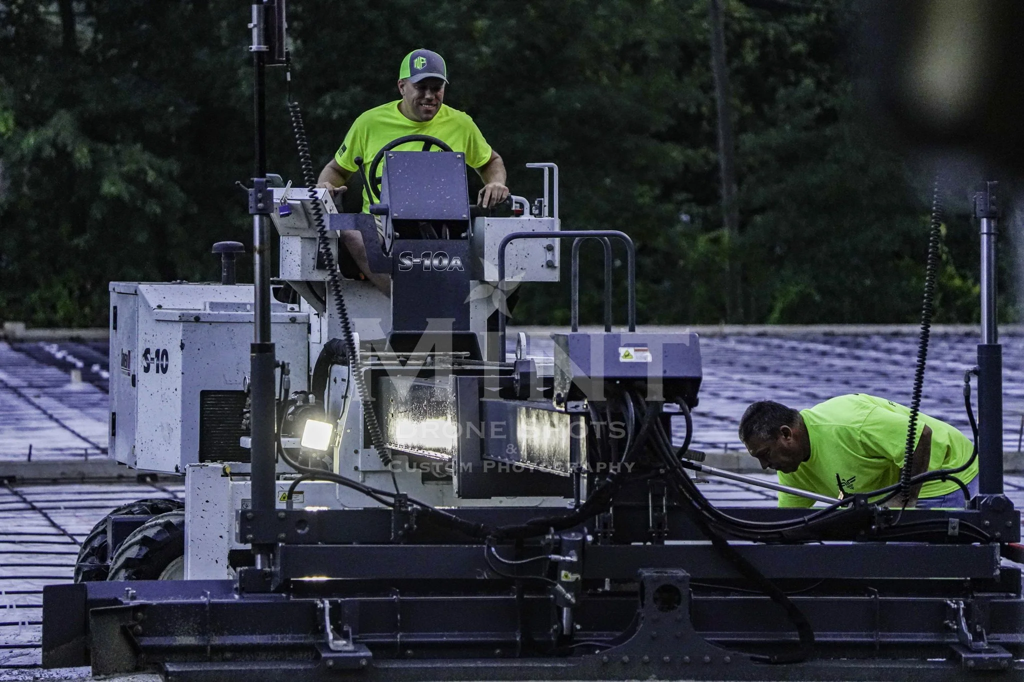 Construction workers operating a machine at a site, with the S-10A model visible, in a wooded area, wearing bright green shirts and a cap.