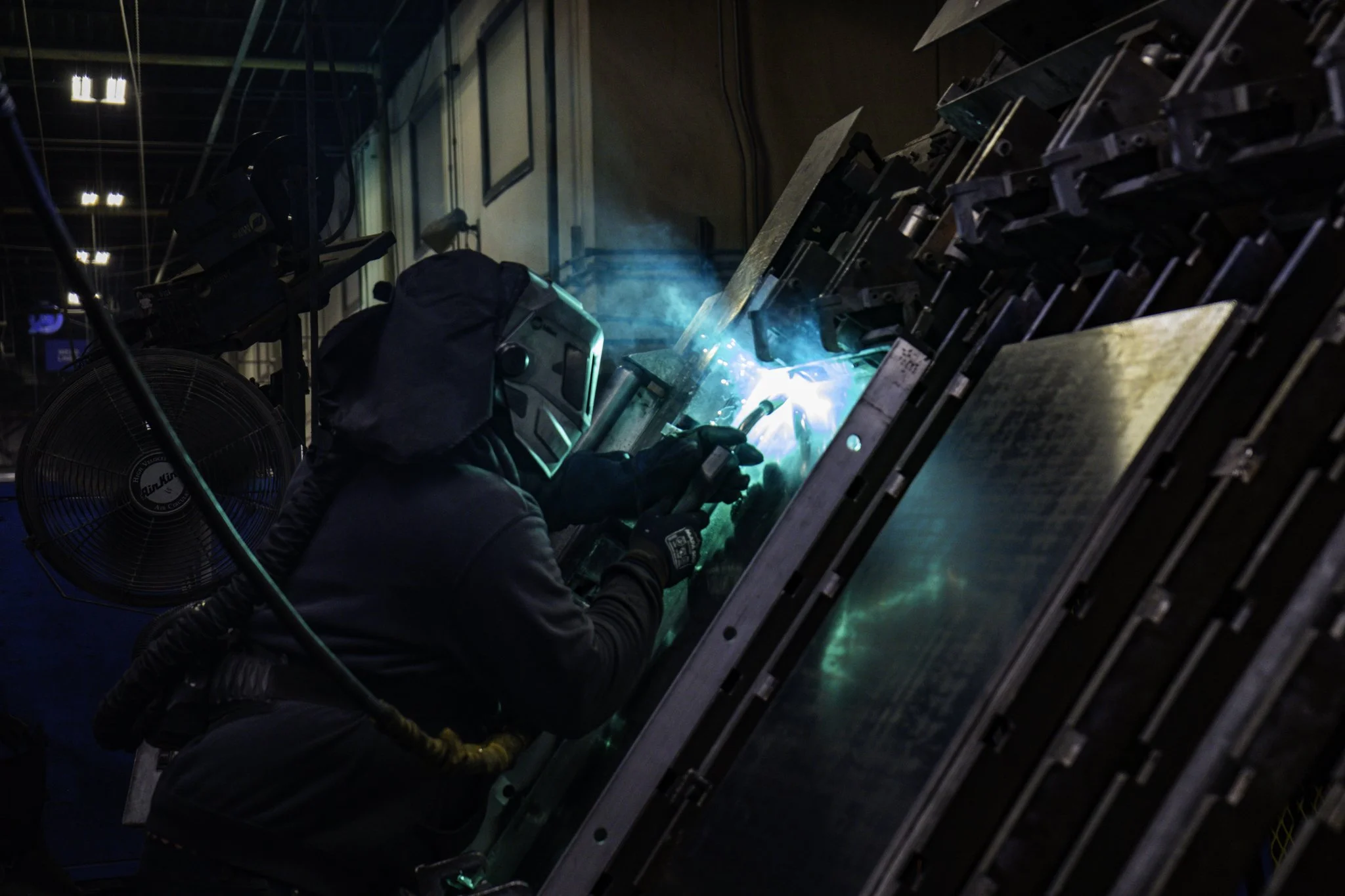 Welder in protective gear working on a large metal structure in a factory setting.