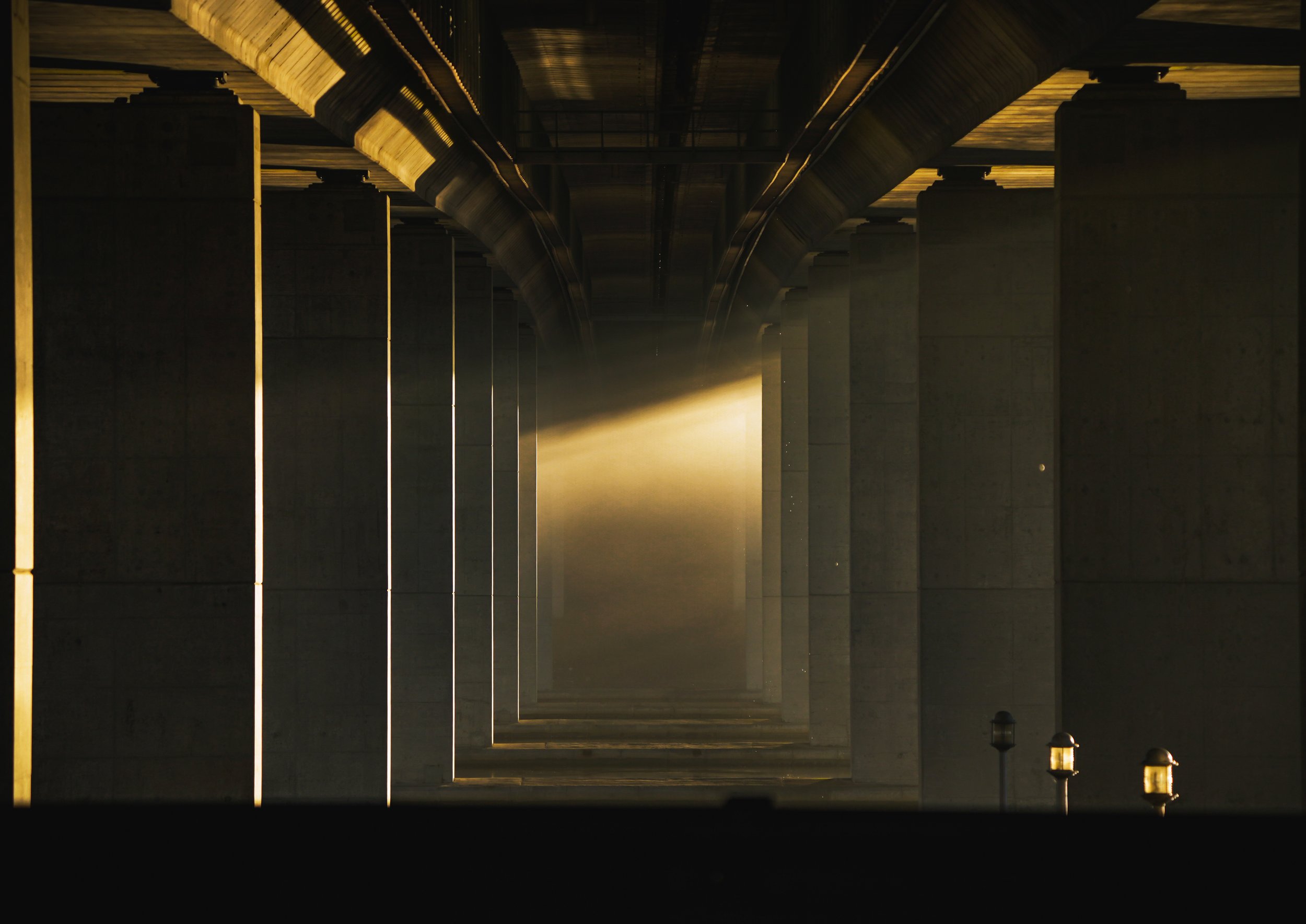 Underneath a bridge with concrete pillars, illuminated by sunlight streaming through, creating dramatic shadows and highlights.