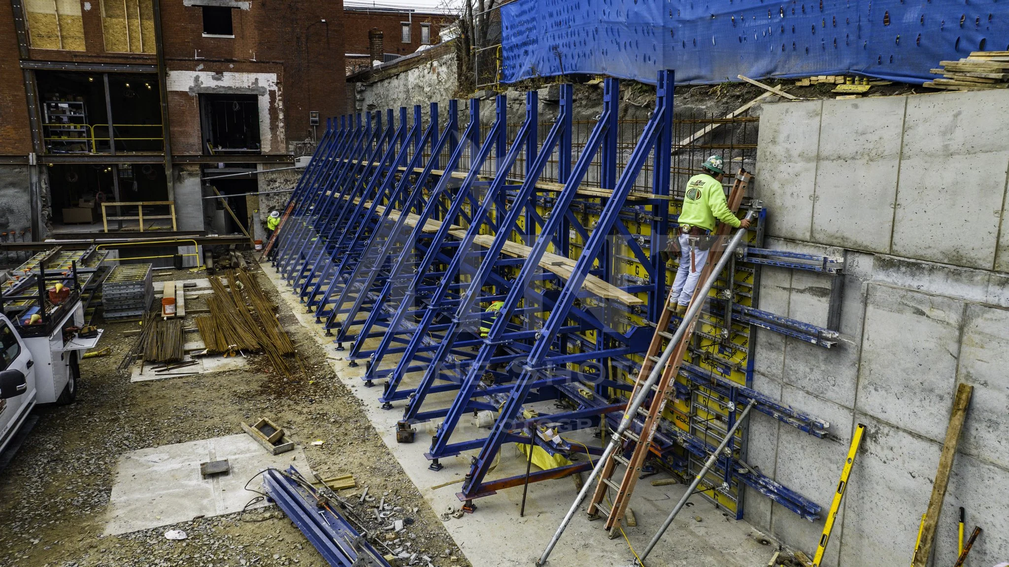 Construction site with workers and blue scaffolding supports against a concrete wall.