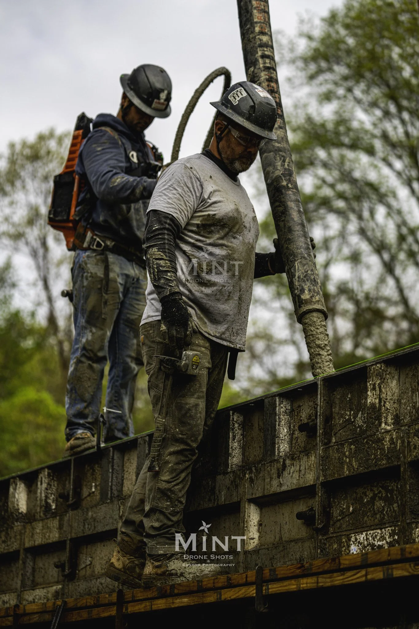Construction workers pouring concrete into a form, wearing hard hats and protective gear, outdoors, with trees in the background.