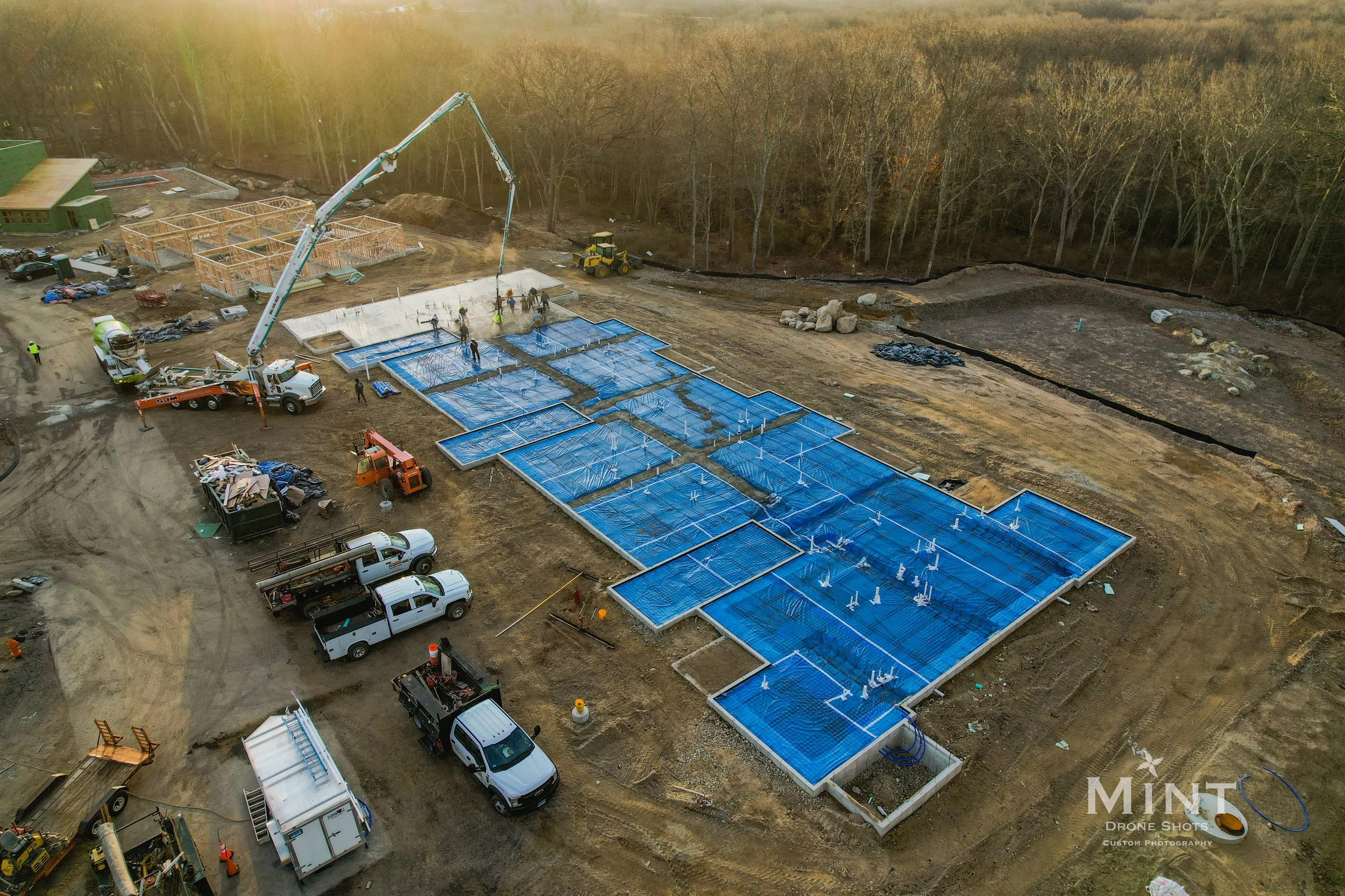 Aerial view of a construction site with concrete being poured by crane onto a prepared foundation. Several trucks and construction vehicles are present, and structures are visible at different stages of assembly. The site is surrounded by bare trees 