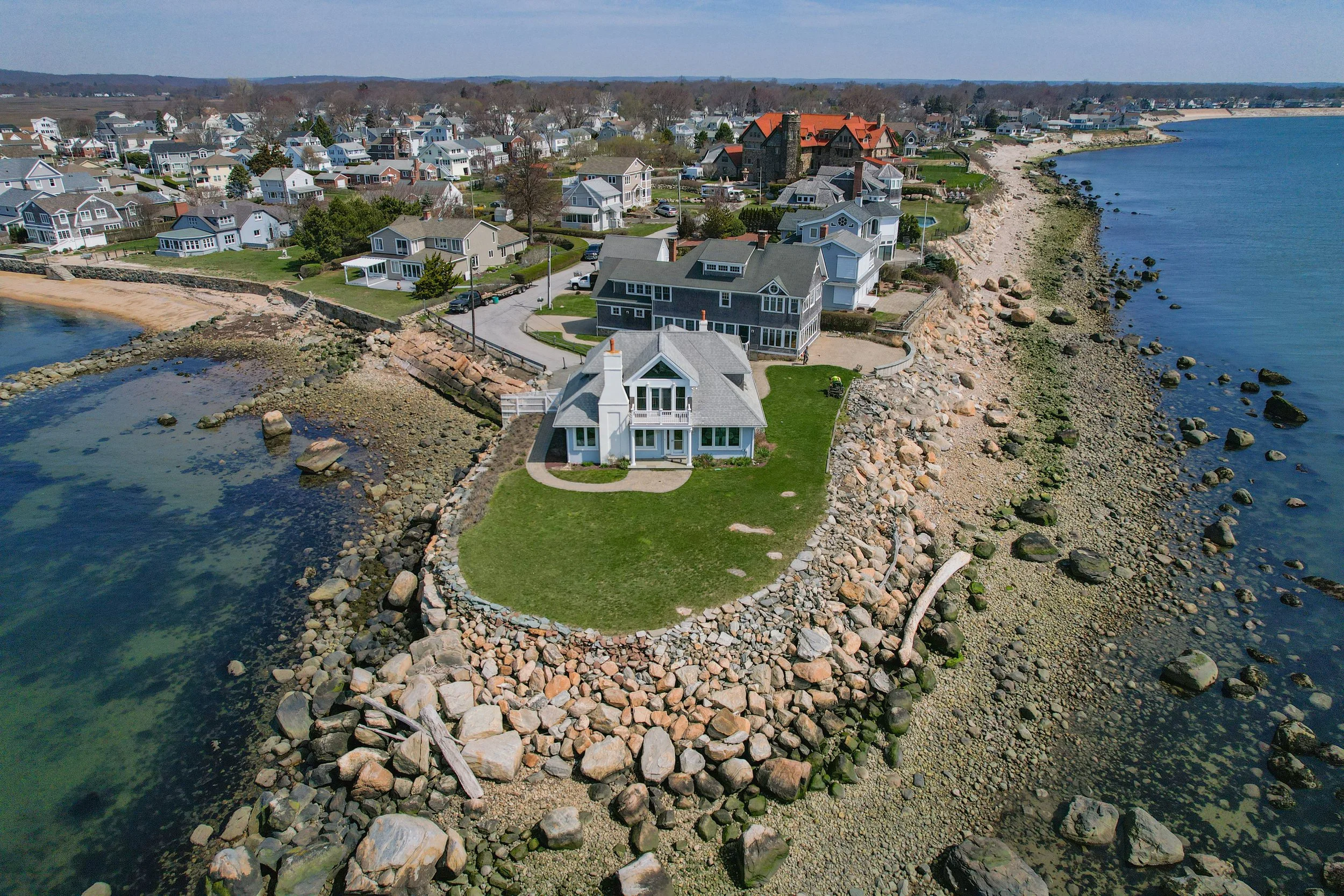 Aerial view of a coastal residential area with houses along a rocky shoreline and adjacent to the ocean, featuring grassy lawns and clear skies.