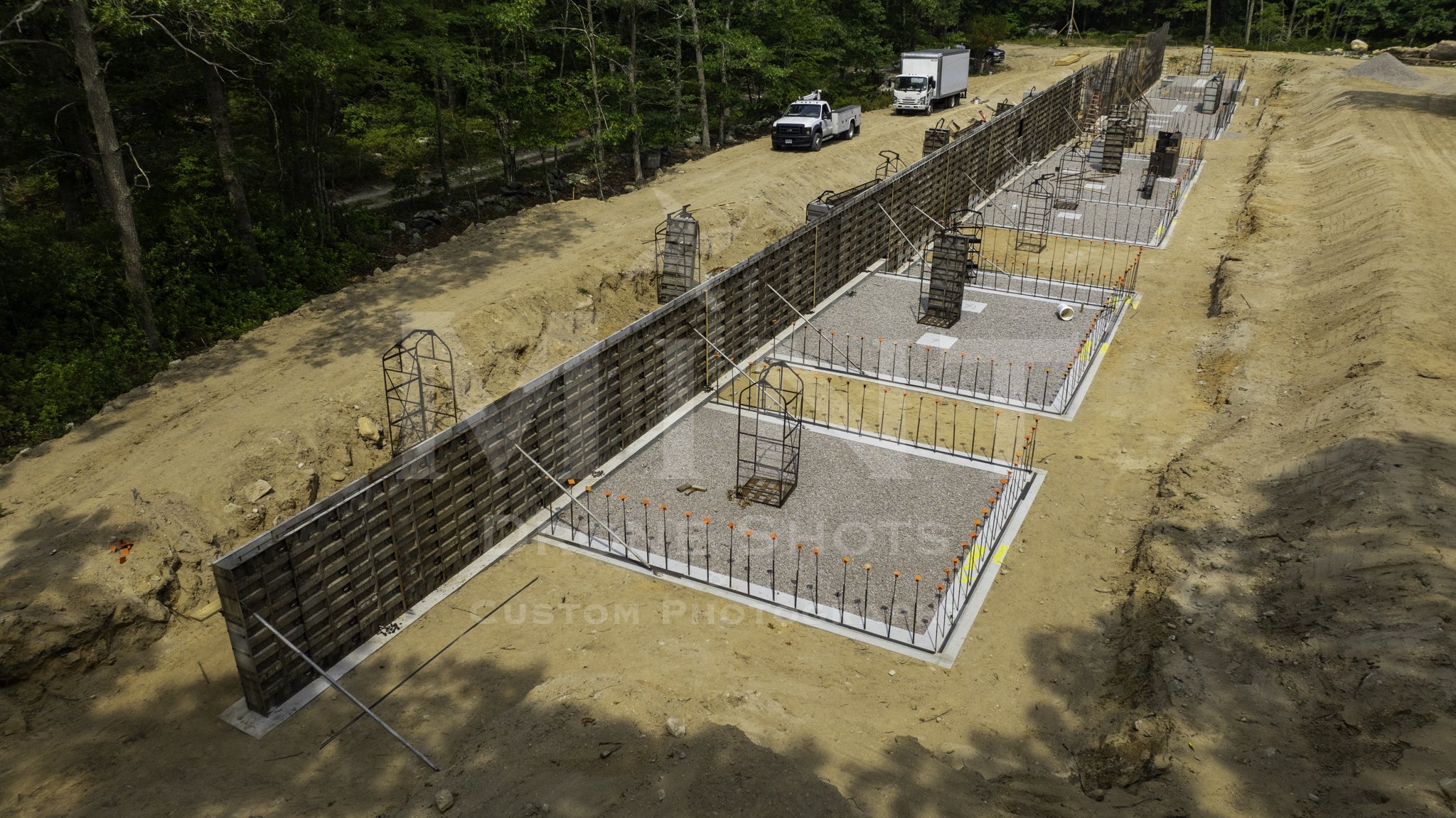 Construction site with concrete foundations and rebar frames in a dug-out area, surrounded by trees, with a truck and a van on a dirt path.