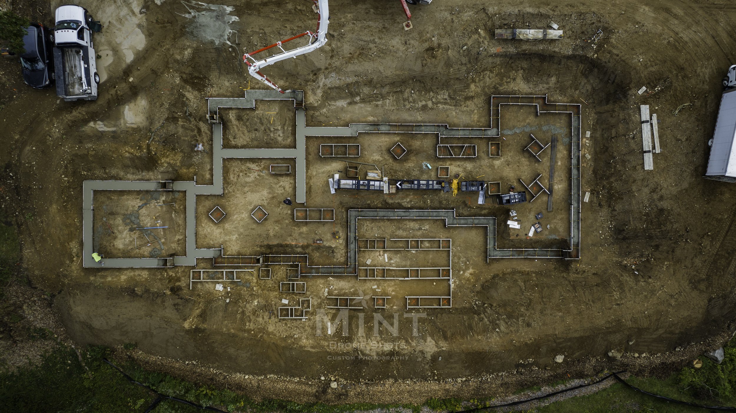 Aerial view of an active construction site with a building foundation layout. Concrete forms and structures are visible on a dirt surface, with construction vehicles and materials present around the site.