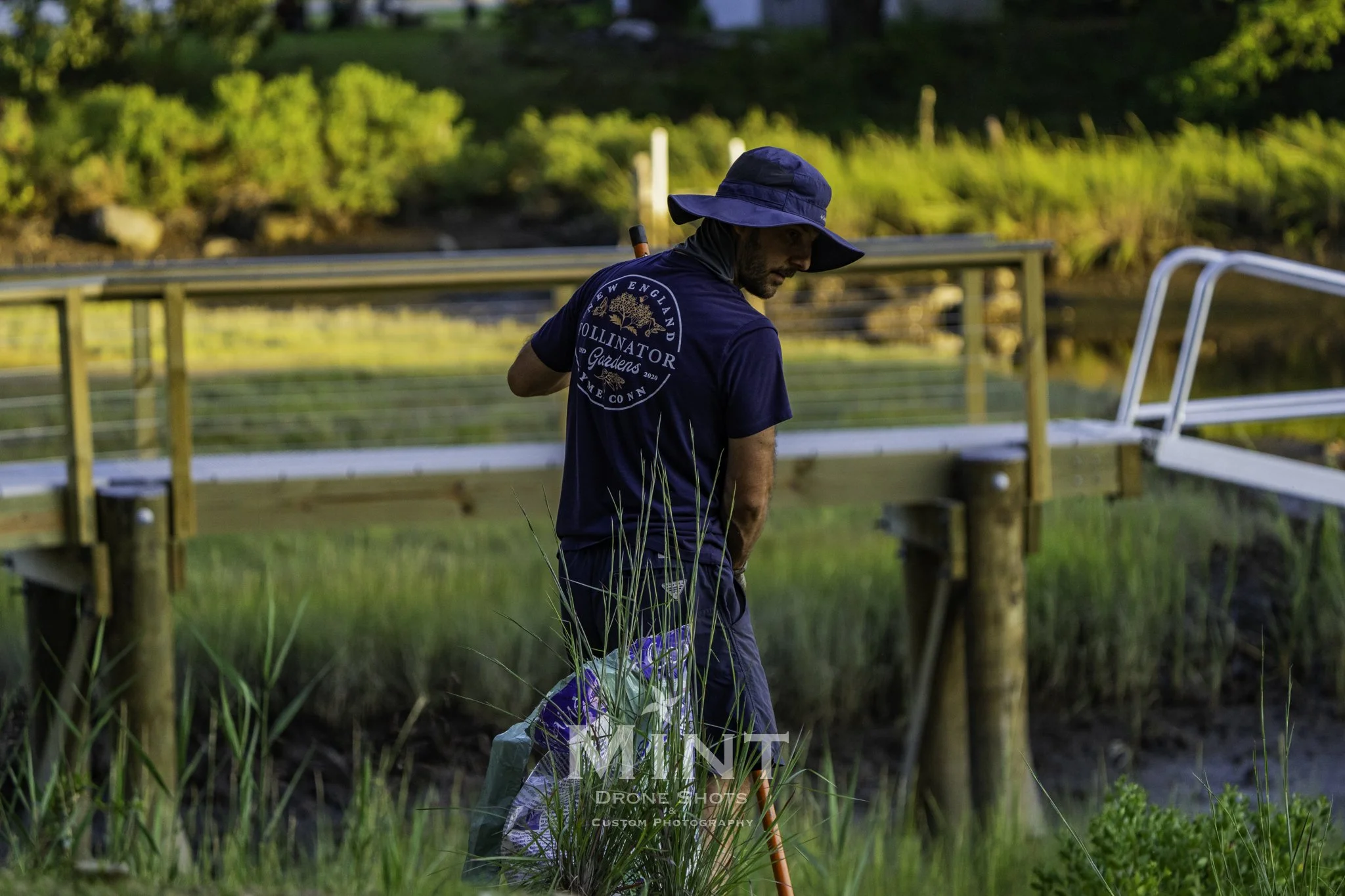 Person wearing a hat and t-shirt with "New England Pollinator Gardens" logo, carrying a watering can on a grassy area near a dock.
