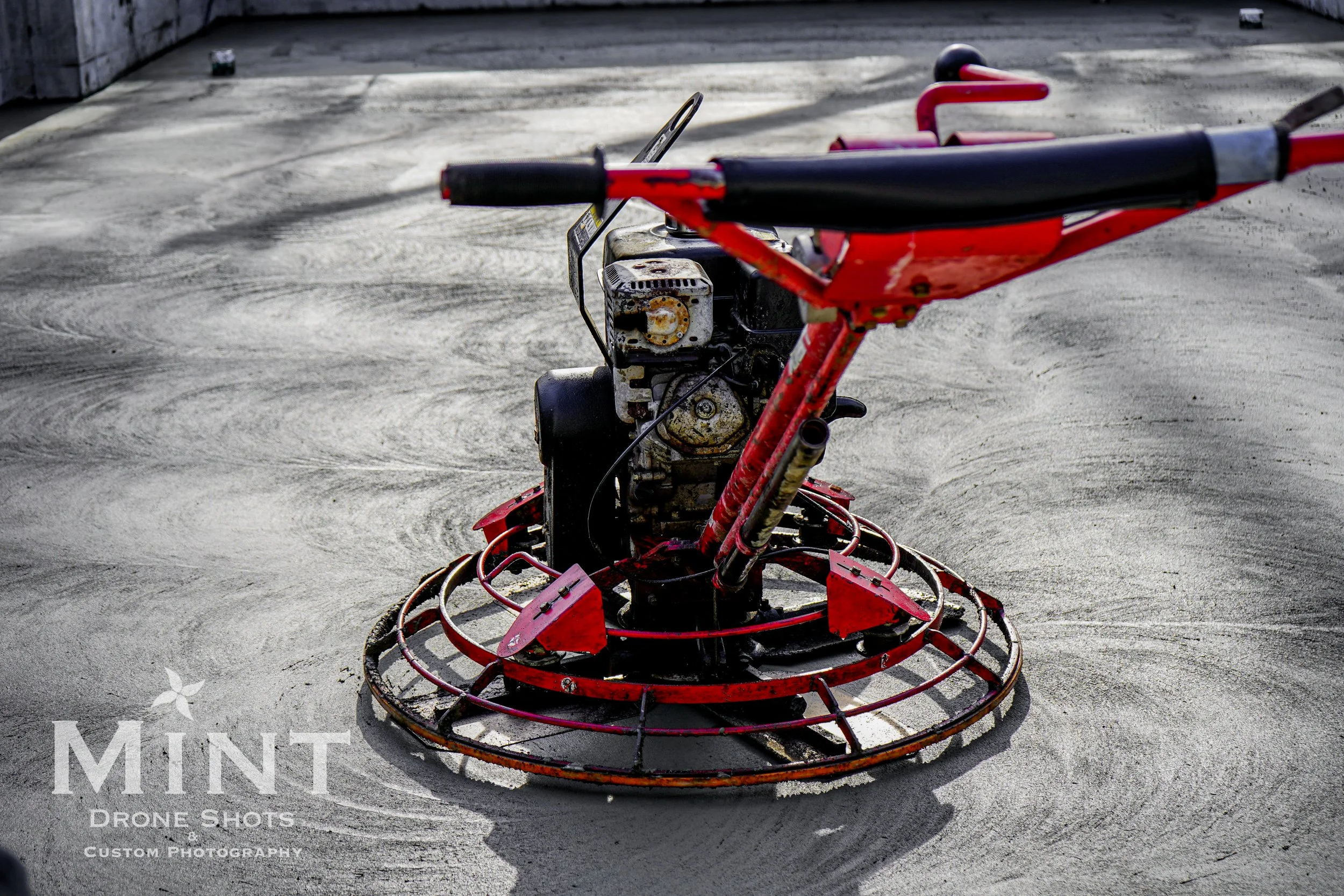 Close-up of a concrete power trowel on a freshly laid cement floor, showing a red machine with metal blades and handle.