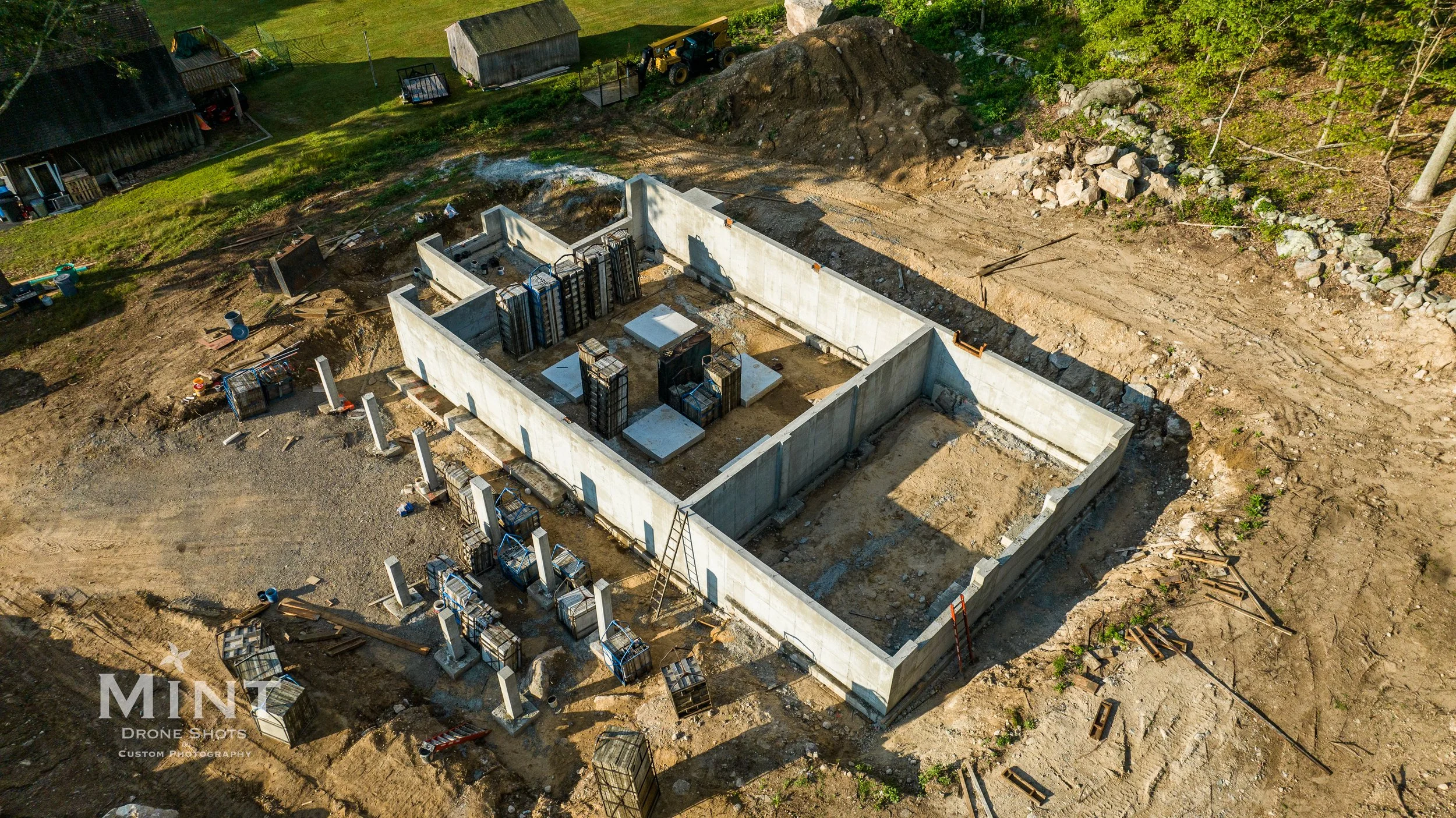 Aerial view of a construction site with concrete foundation walls, surrounding open land, construction materials, and machinery.