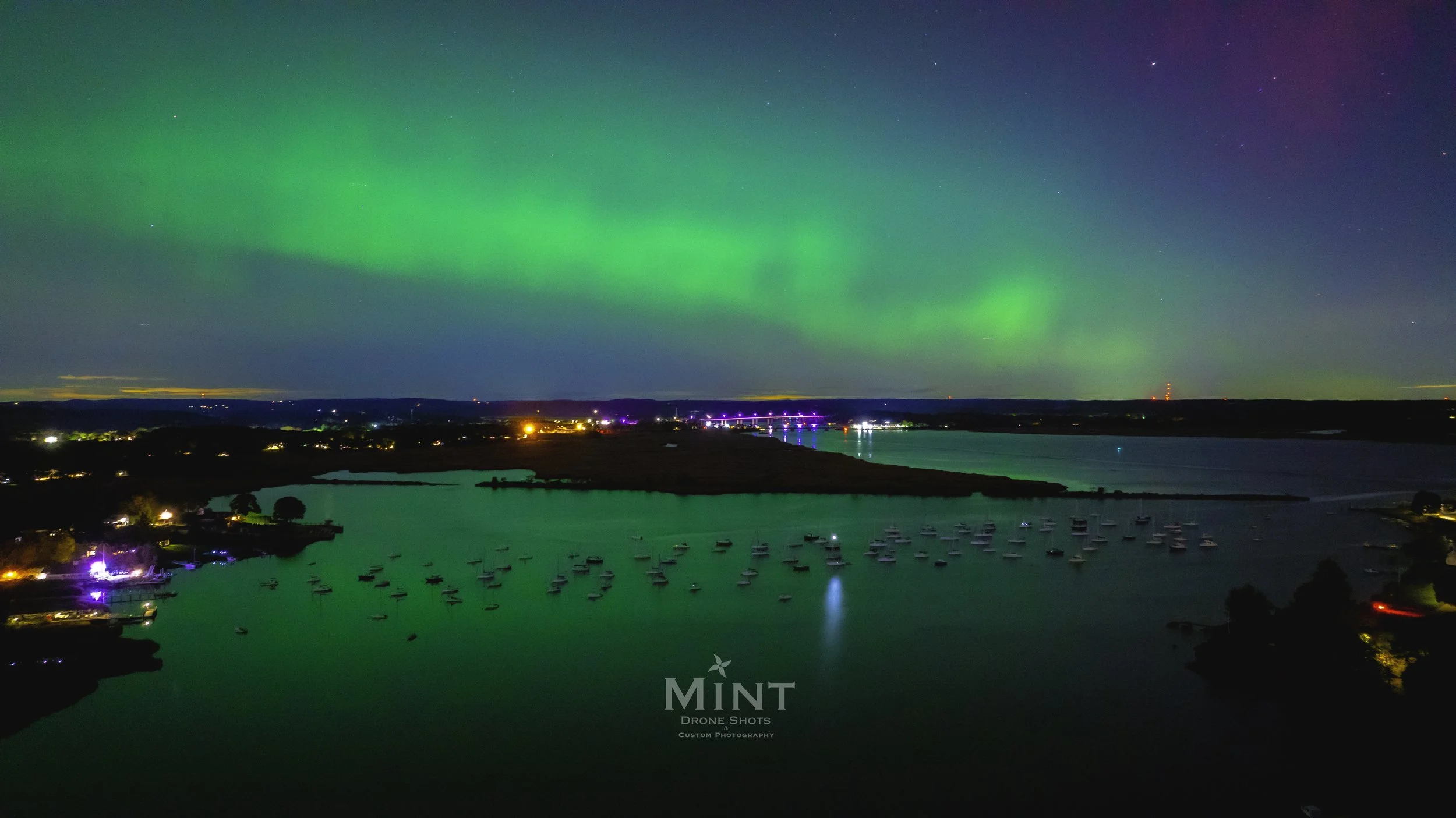 Northern lights over a waterfront with anchored boats at night, colorful city lights and reflections on the water.