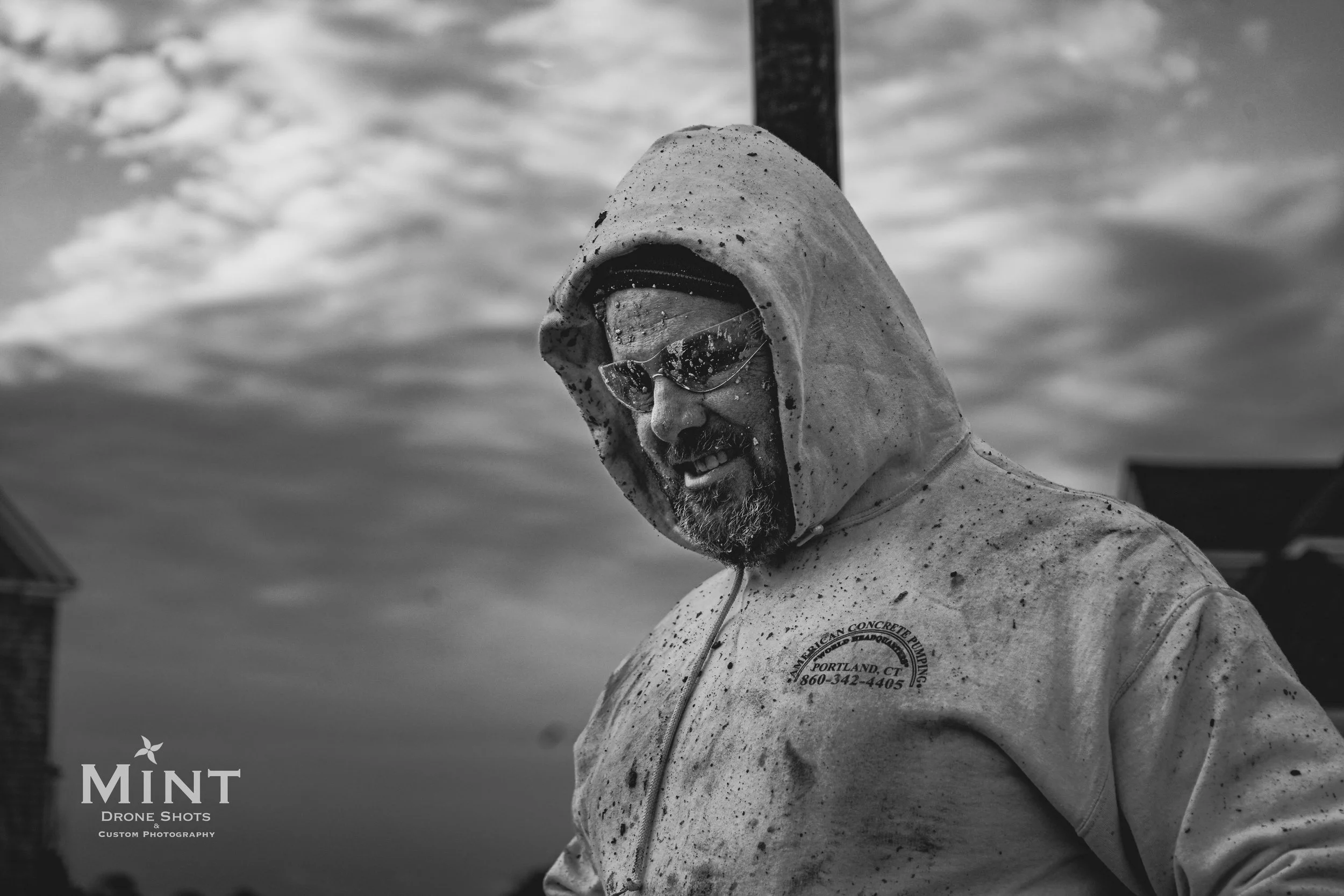 Black and white photo of a person in a hooded sweatshirt and protective glasses, working outdoors, with a cloudy sky background.