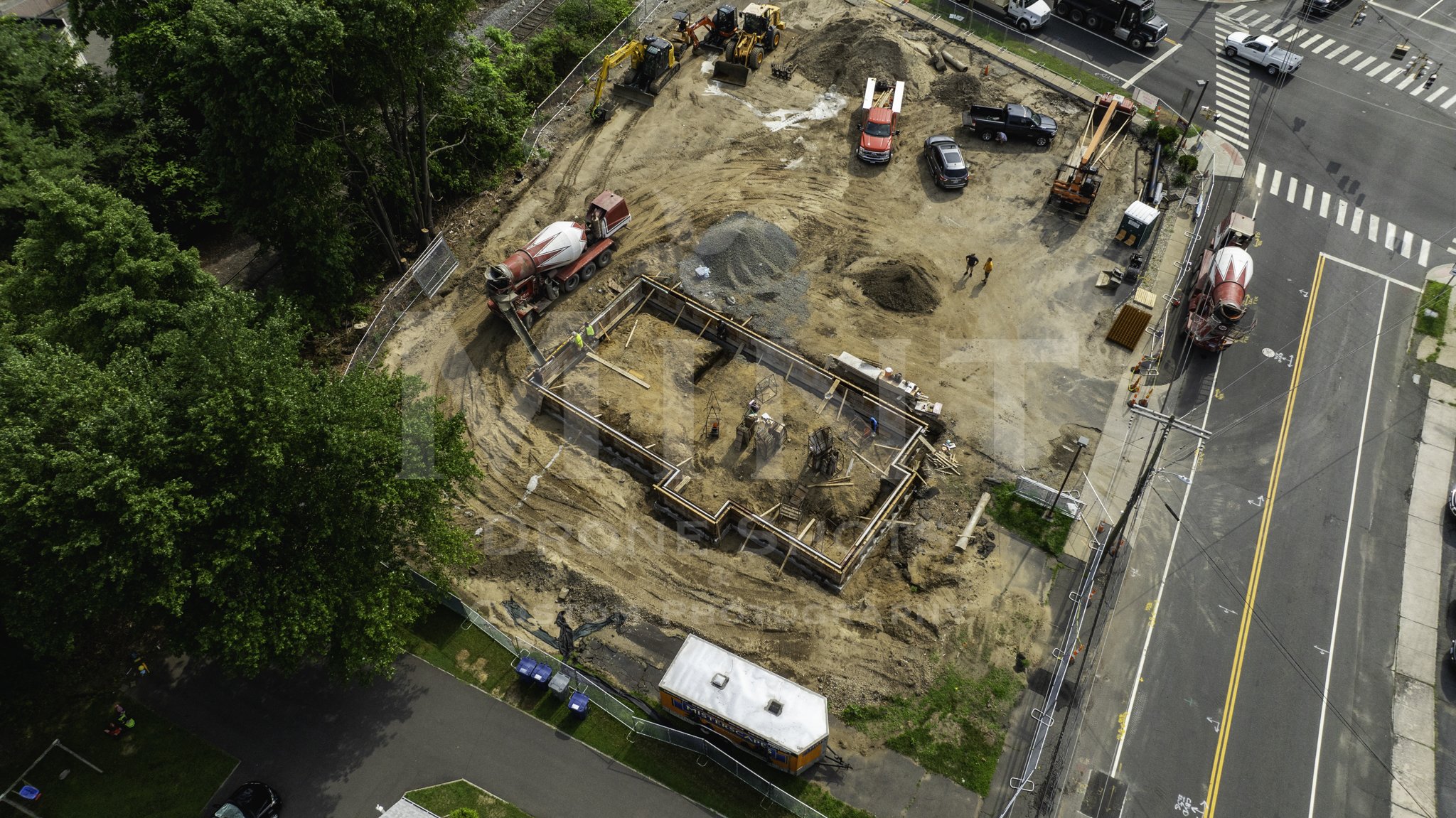 Aerial view of a construction site with machinery, trucks, and workers. Foundations are being laid out on a dirt area surrounded by trees and adjacent to a road intersection.