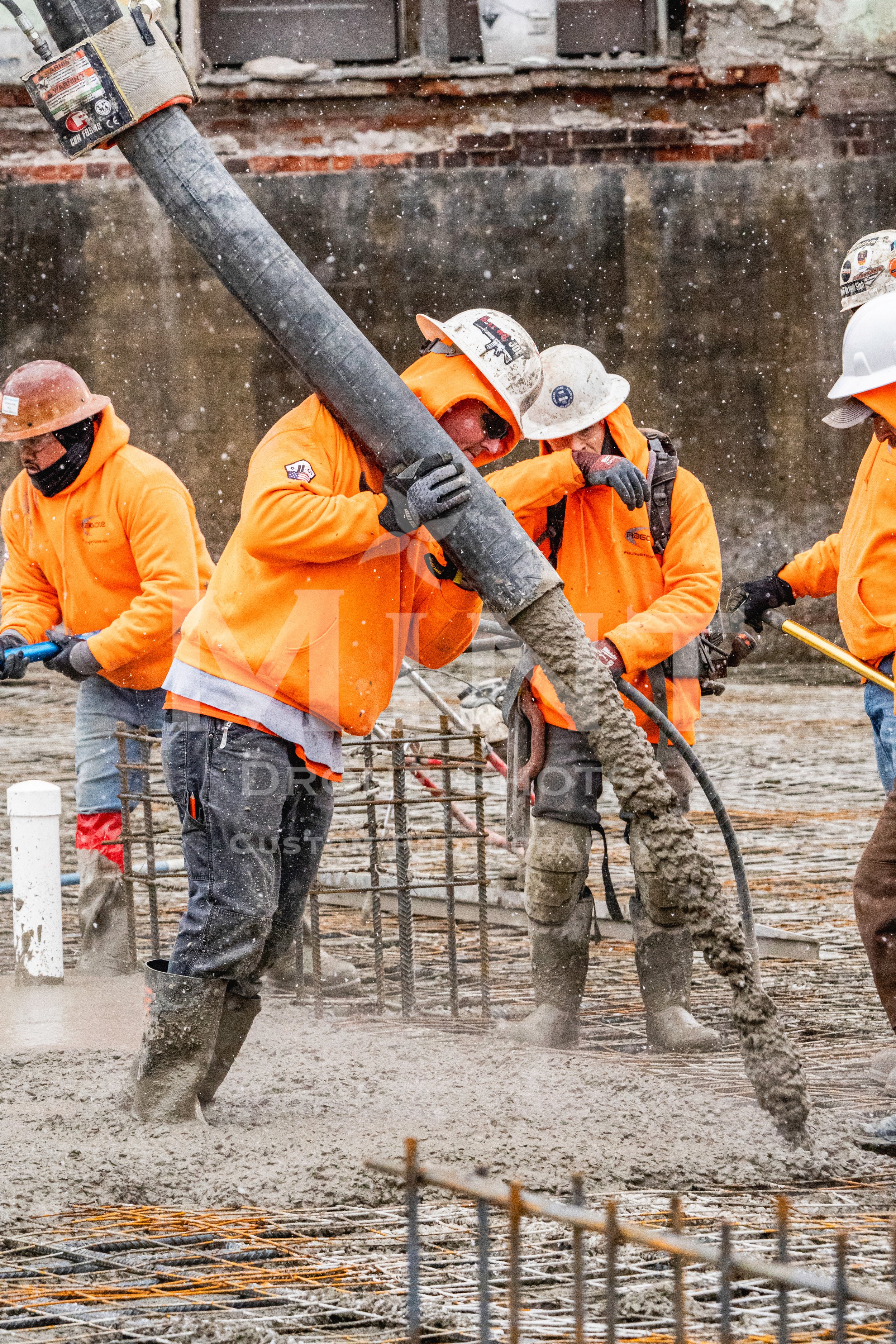 Construction workers pouring concrete at a construction site, wearing orange safety jackets and hard hats.