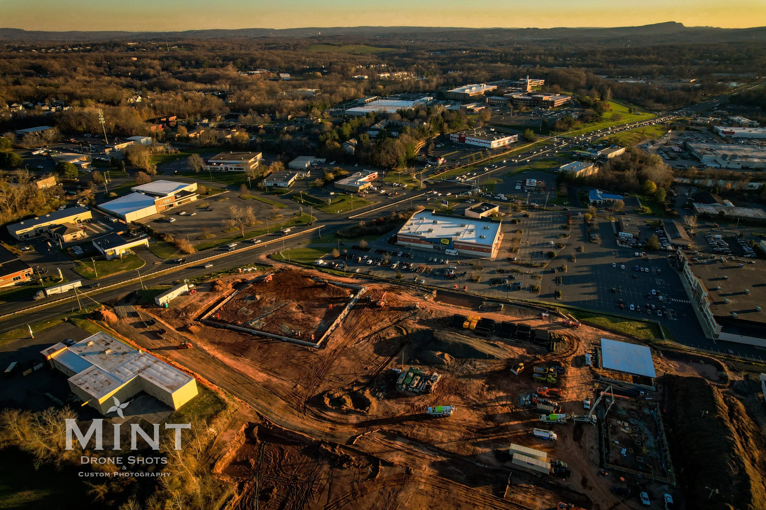 Aerial view of a construction site and surrounding urban area with roads, commercial buildings, and parked cars.