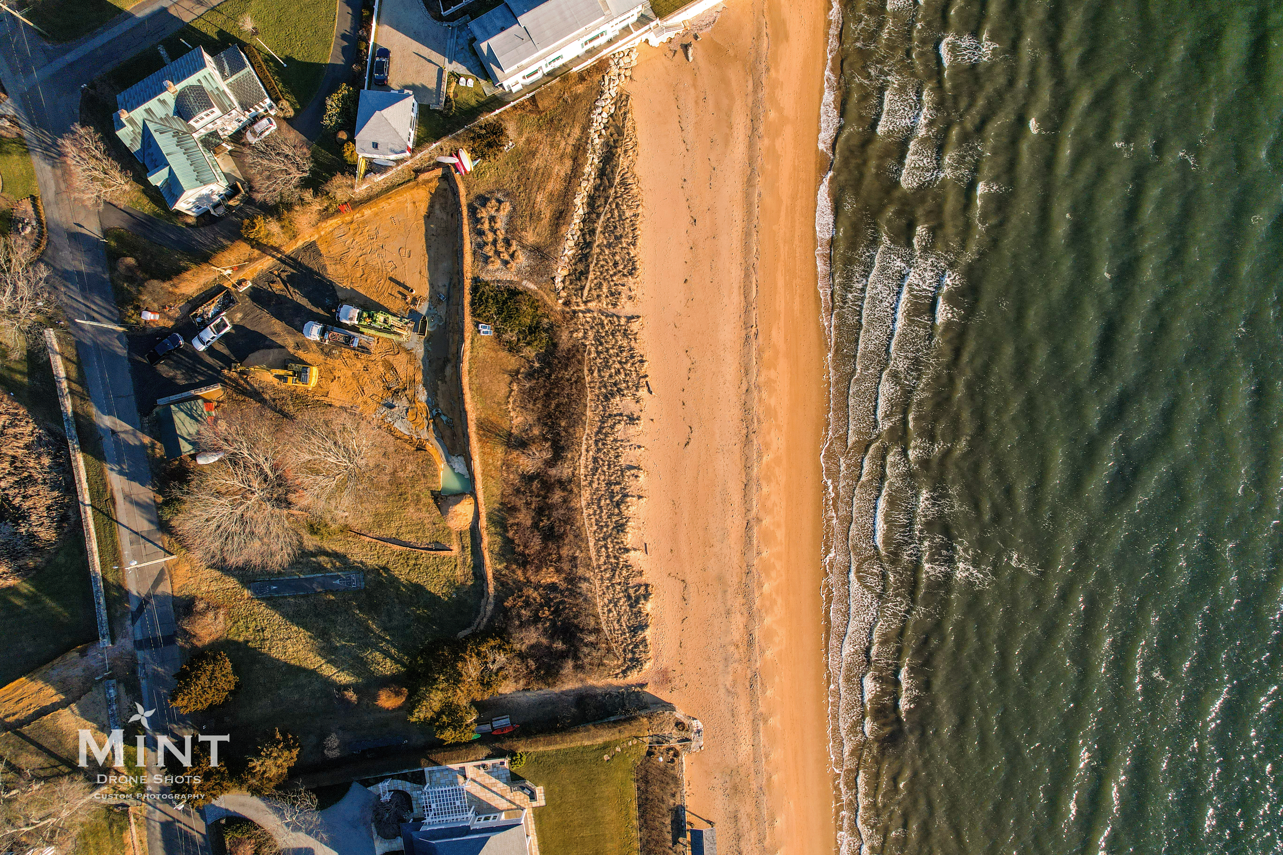 Aerial view of a beachfront area with houses, construction vehicles, and equipment near the sand and ocean.