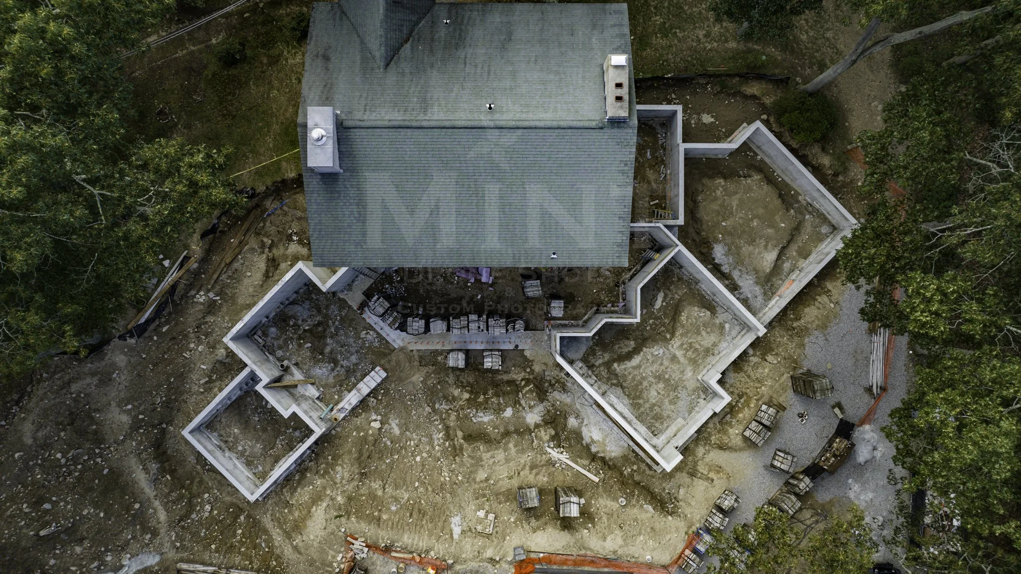 Aerial view of a construction site with a partially constructed building surrounded by trees. The site shows foundational work, including concrete walls and organized construction materials.