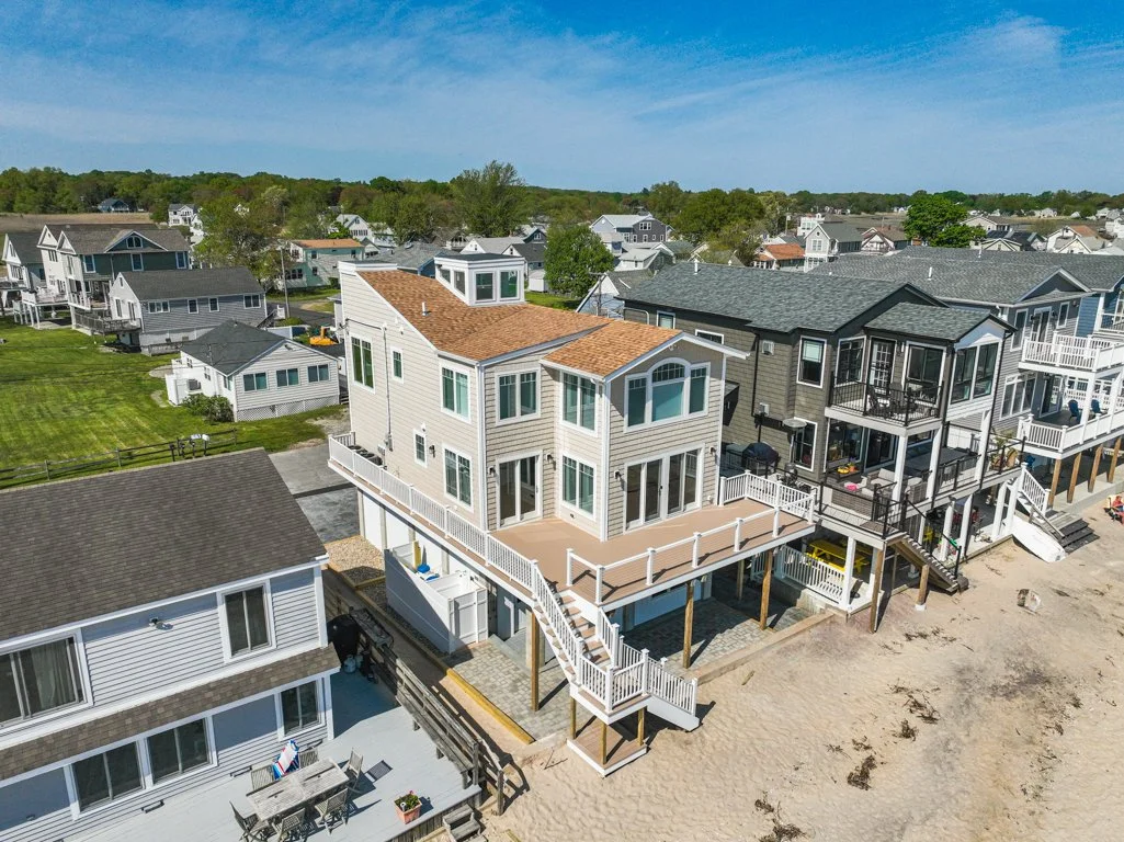 Aerial view of a beachfront house with a wooden deck and stairs leading to the sand, surrounded by other coastal homes and greenery under a clear blue sky.