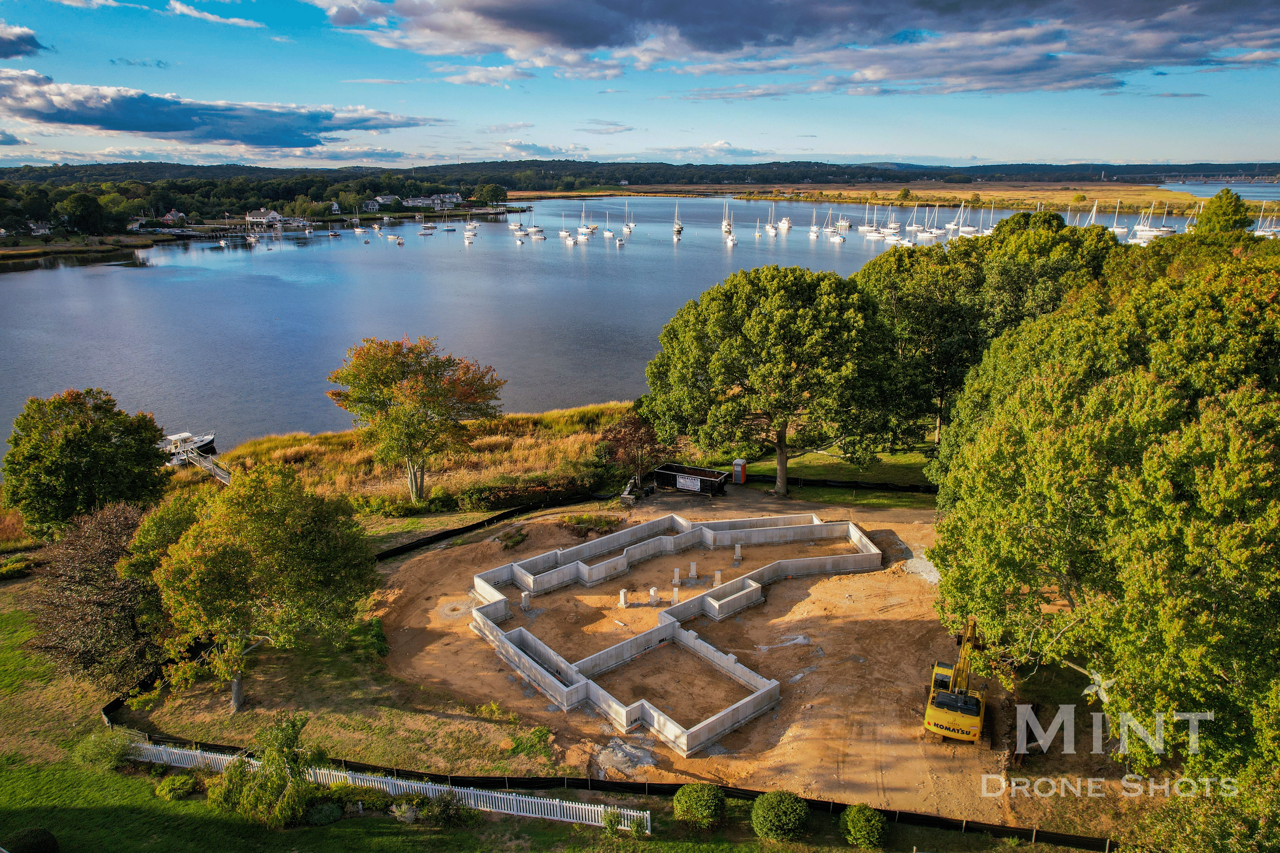 Drone view of a scenic waterfront construction site with a house foundation, surrounded by trees and near a body of water with boats.