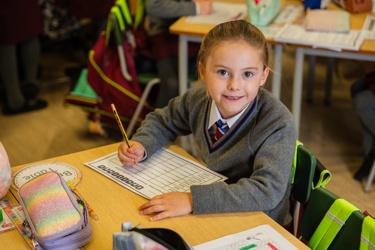 A girl in a grey jumper and tie looks up from a spelling worksheet she is filling in with a pencil