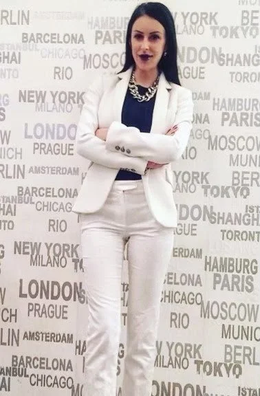 A woman with black hair wearing a white suit with a black top and pearl necklace, standing with arms crossed in front of a wall with city names in black and gray text.