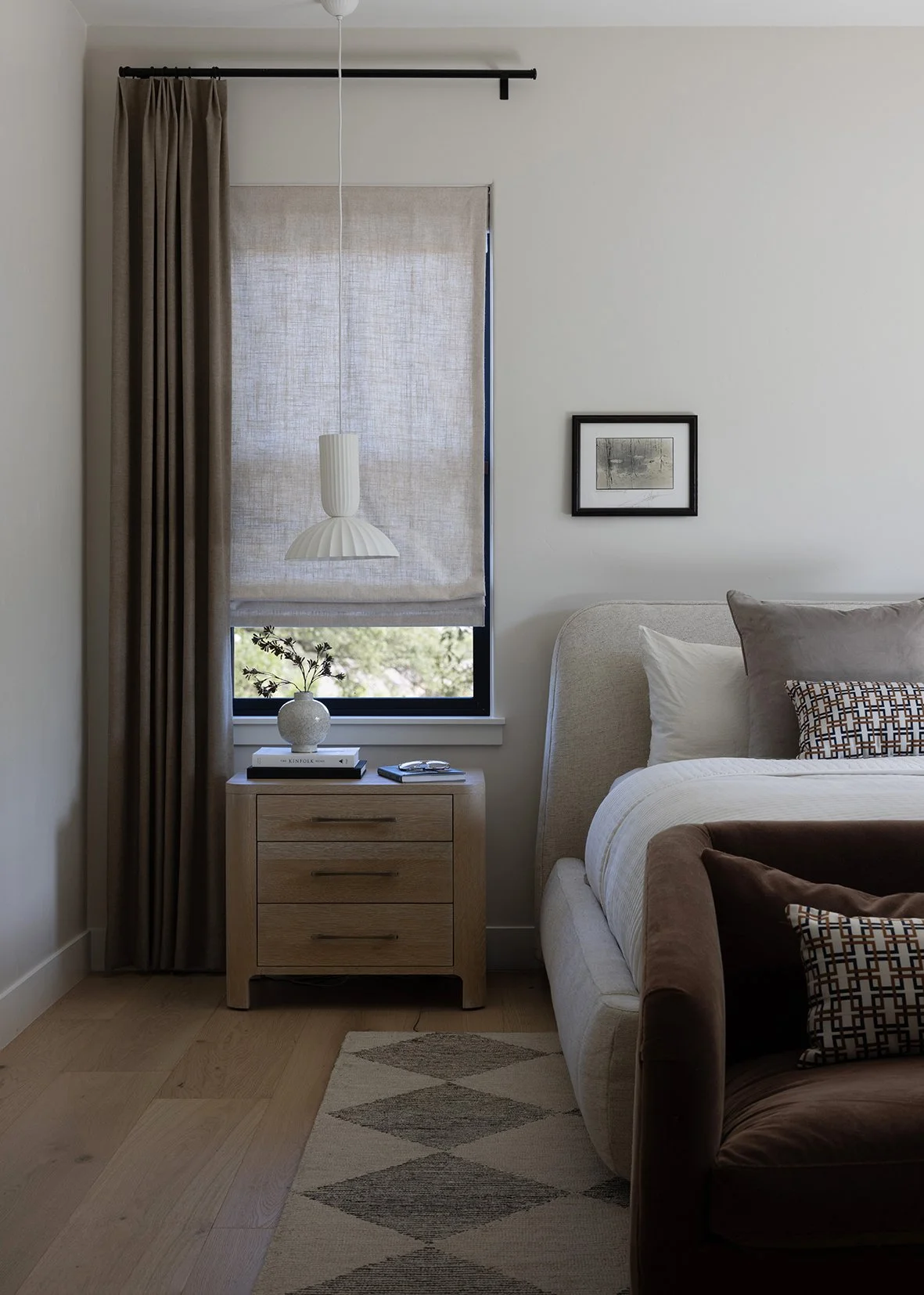 This serene bedroom design combines soft textures, warm wood tones, and clean lines to create a calm, restful retreat. A custom oak nightstand and layered window treatments add warmth and functionality, while tailored upholstery, neutral bedding, and