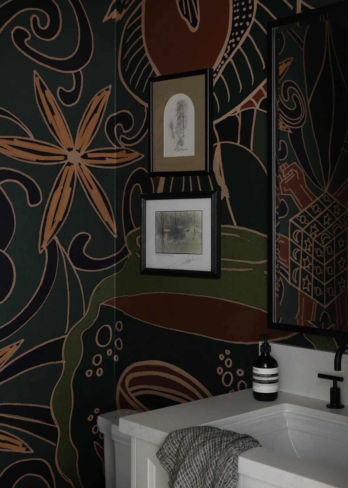 This custom powder room makes a bold statement with richly patterned wallpaper and layered, moody tones that instantly elevate the space. The oversized botanical mural wraps the walls in deep greens, rusts, and warm neutrals, creating a dramatic back