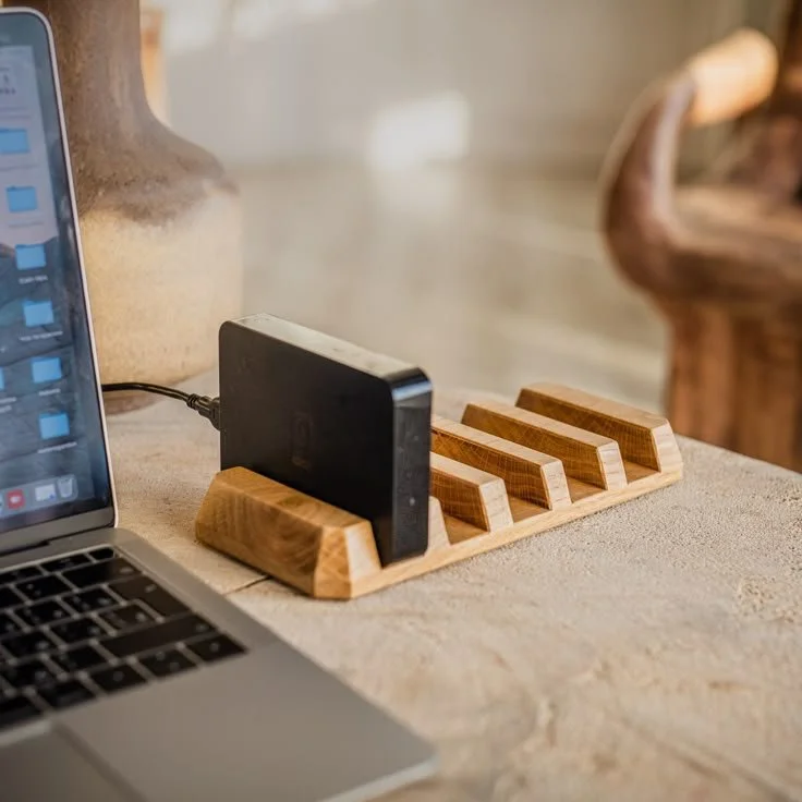A wooden phone stand on a beige surface with a black smartphone placed on it, charging via a cable. Part of a laptop is visible on the left, and a blurred background with a vase and furniture.
