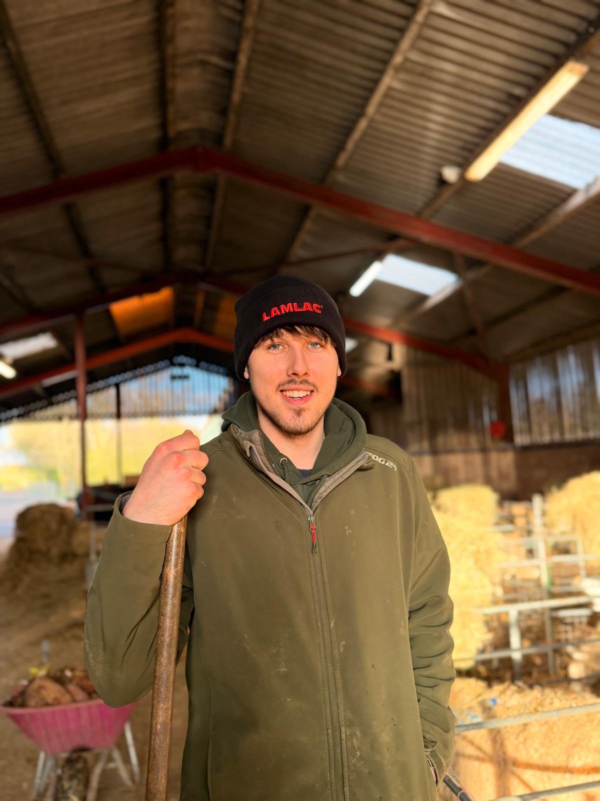 Young man in a barn or stable with hay bales, wearing a black hat with red text, green jacket, holding a pitchfork, smiling, with wooden walls and a sloped metal roof.