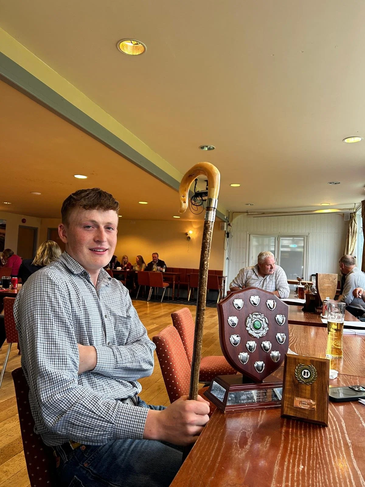 A young man in a checked shirt sits at a restaurant table holding a walking stick with a curved handle, next to a shield-shaped trophy with smaller shields around a central emblem, and a glass of beer. Other patrons are seated at tables in the background.