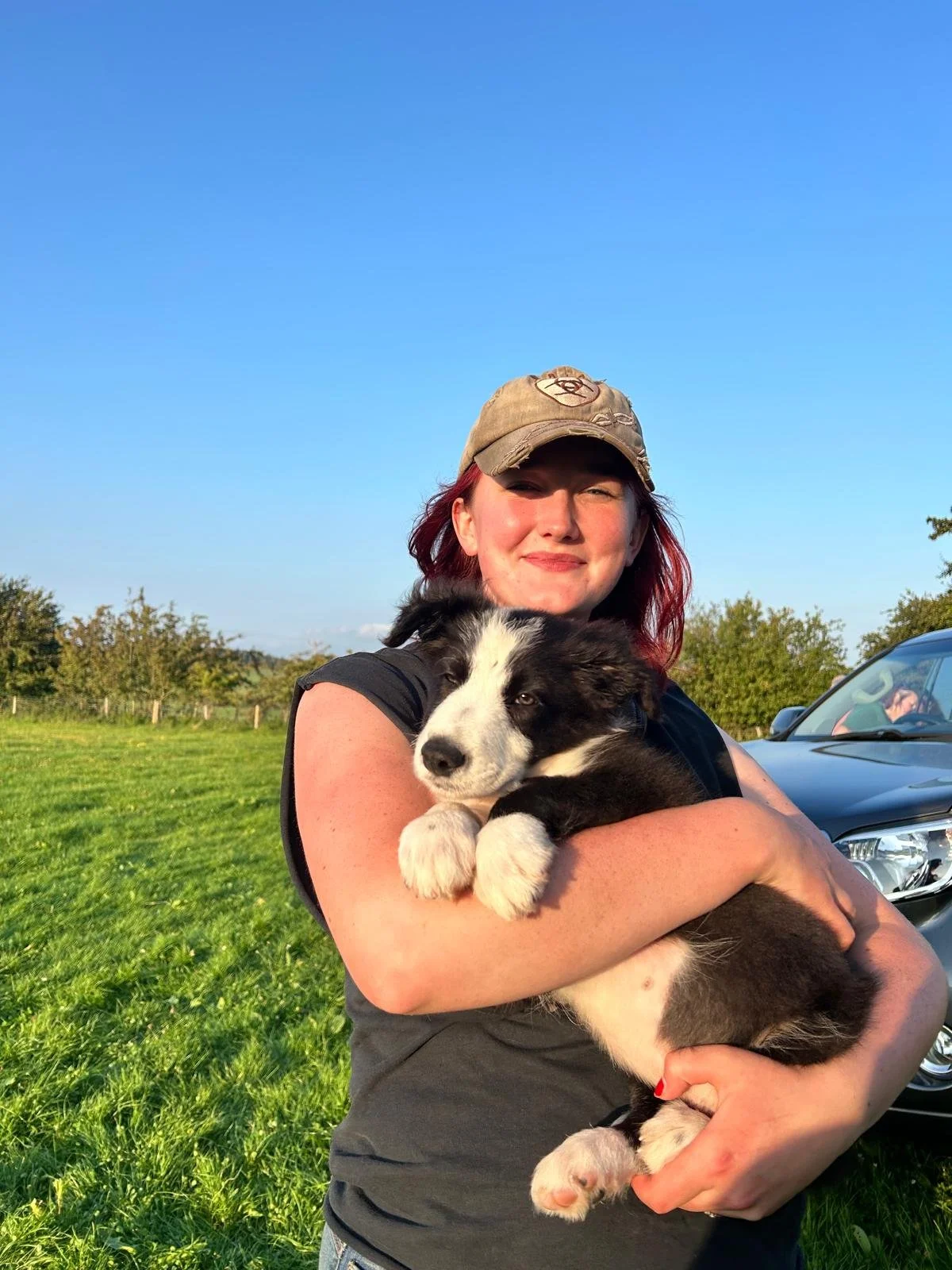Woman holding a black and white puppy outdoors on a sunny day with greenery and a parked car in the background.